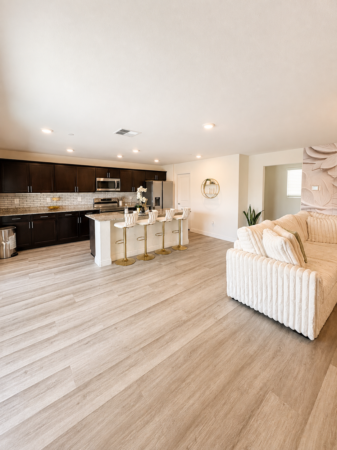 A modern kitchen with dark brown cabinets, stainless steel appliances, a granite countertop island with four white and gold bar stools, gray wood flooring, a white wall with a round wall shelf, and a potted plant.
