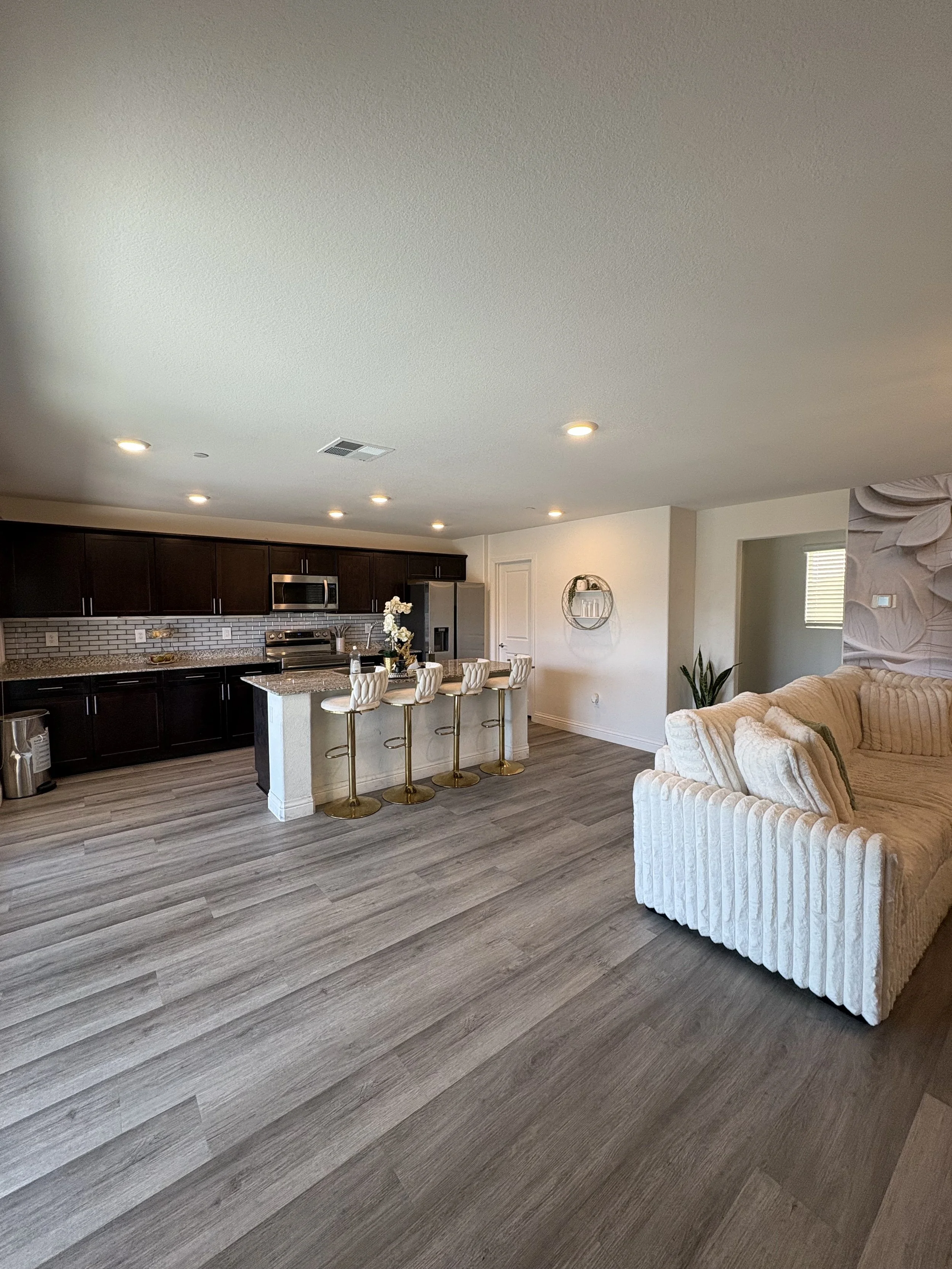 A modern kitchen with dark brown cabinets, stainless steel appliances, a granite countertop island with four white and gold bar stools, gray wood flooring, a white wall with a round wall shelf, and a potted plant.