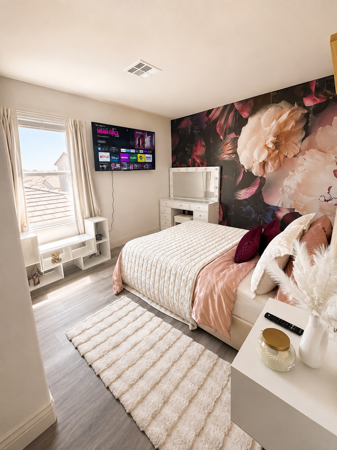 A neatly made bed with beige, cream, and plum-colored bedding and pillows. A white floating nightstand beside it holds a white vase with dried pampas grass and a few small decorative items. There is a framed picture of a perfume bottle and flowers hanging on the plain white wall above the bed.