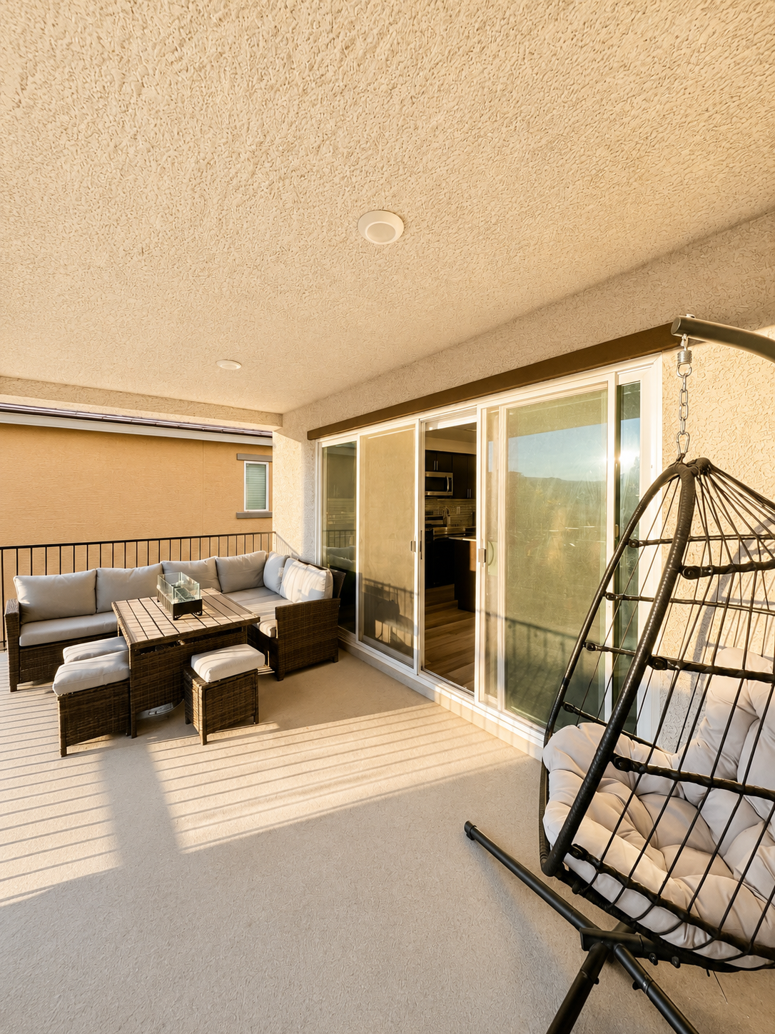 A balcony with outdoor furniture including a sectional sofa, coffee table, and two ottomans, enclosed by a railing, with a sliding glass door leading inside and a hanging egg chair, illuminated by warm sunlight.
