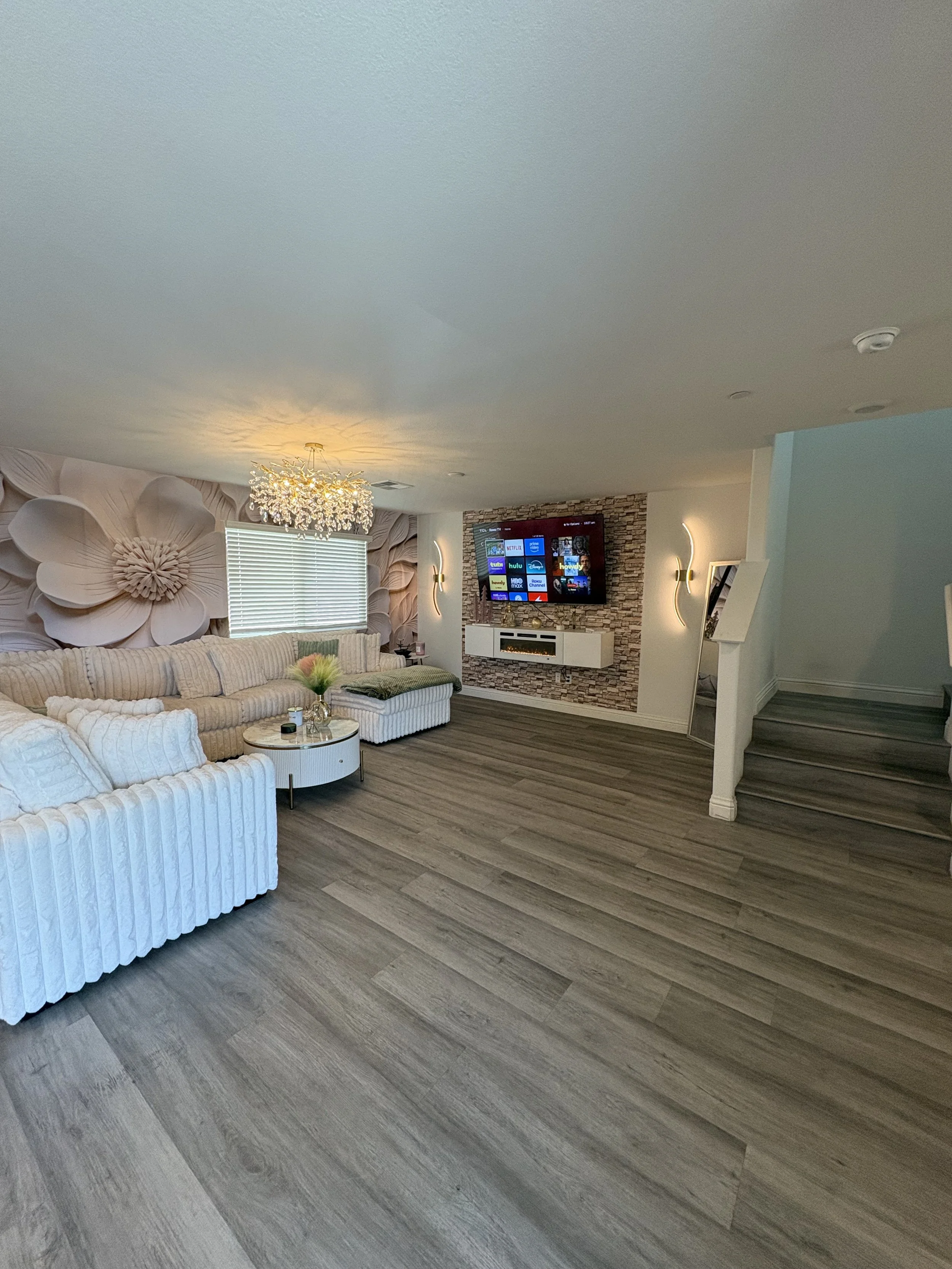 Living room with white striped sofas, a round coffee table, a large window, decorative wall art, a TV mounted on a stone accent wall, a potted plant, a cat lying on a cushion, and wooden flooring.