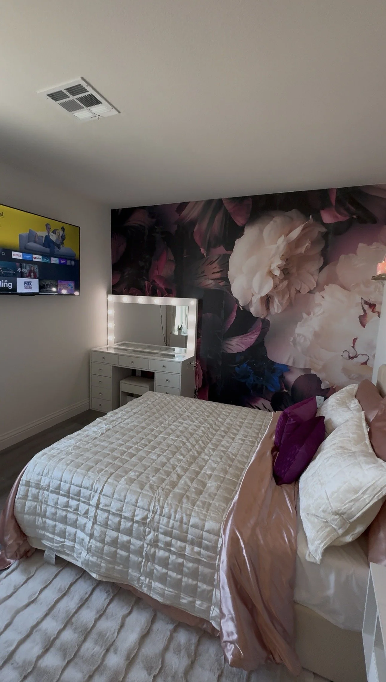 A neatly made bed with beige, cream, and plum-colored bedding and pillows. A white floating nightstand beside it holds a white vase with dried pampas grass and a few small decorative items. There is a framed picture of a perfume bottle and flowers hanging on the plain white wall above the bed.
