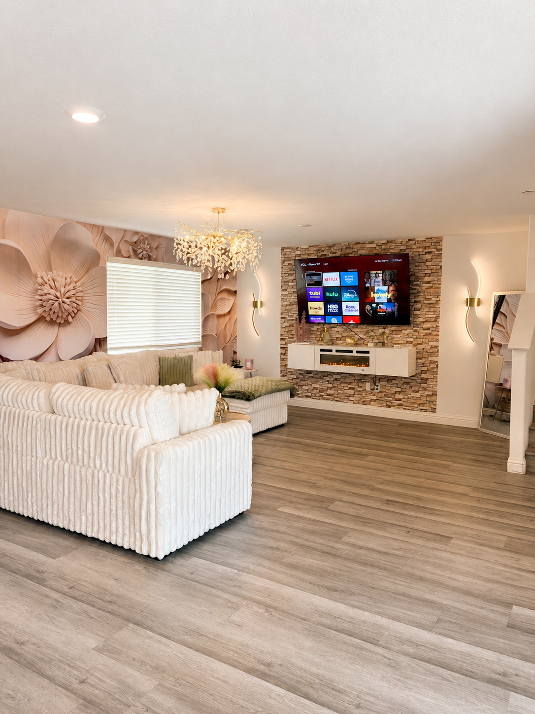 Living room with white striped sofas, a round coffee table, a large window, decorative wall art, a TV mounted on a stone accent wall, a potted plant, a cat lying on a cushion, and wooden flooring.