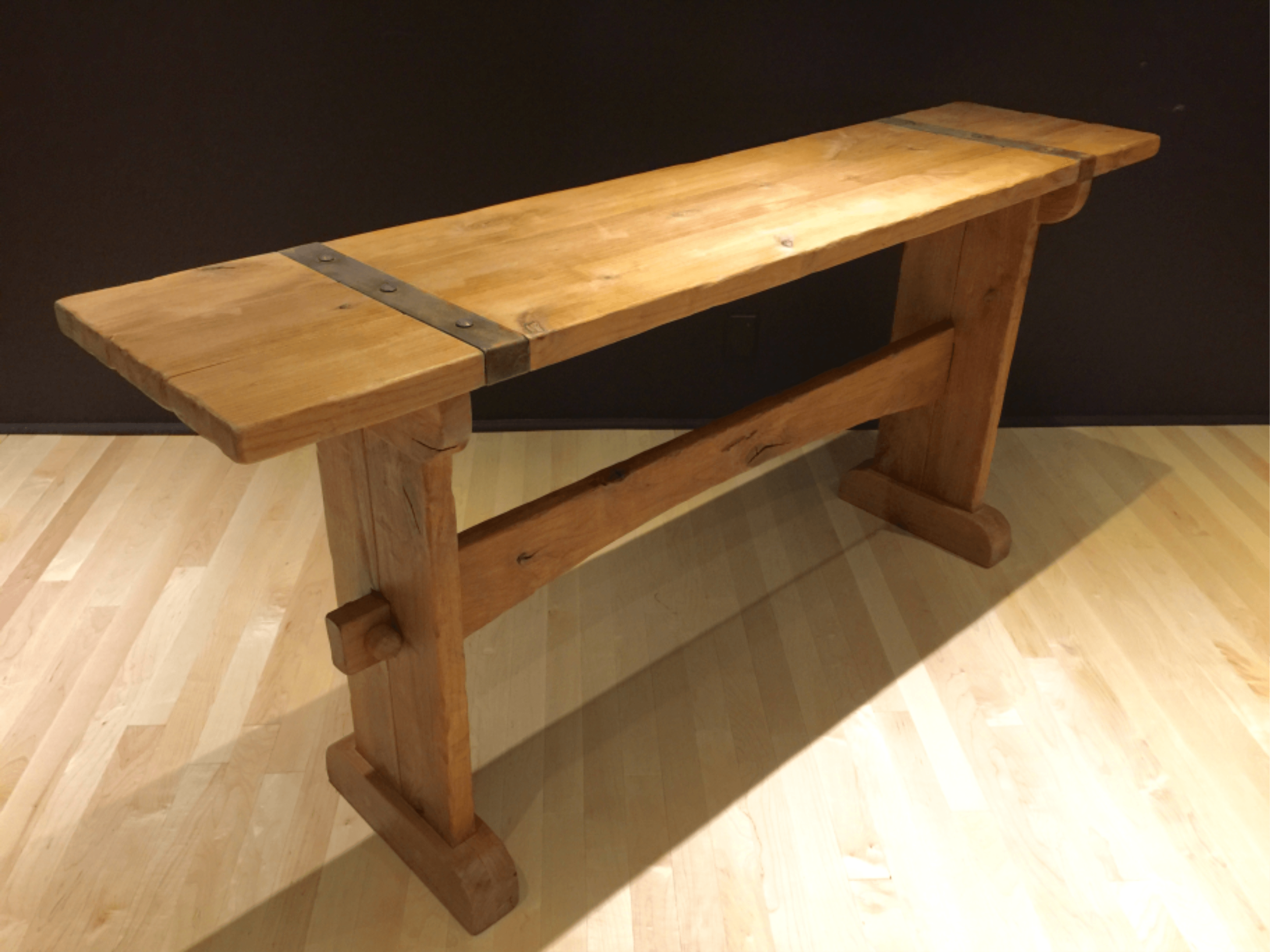 A wooden bench with a rustic design against a dark background, featuring metal brackets on the top surface.