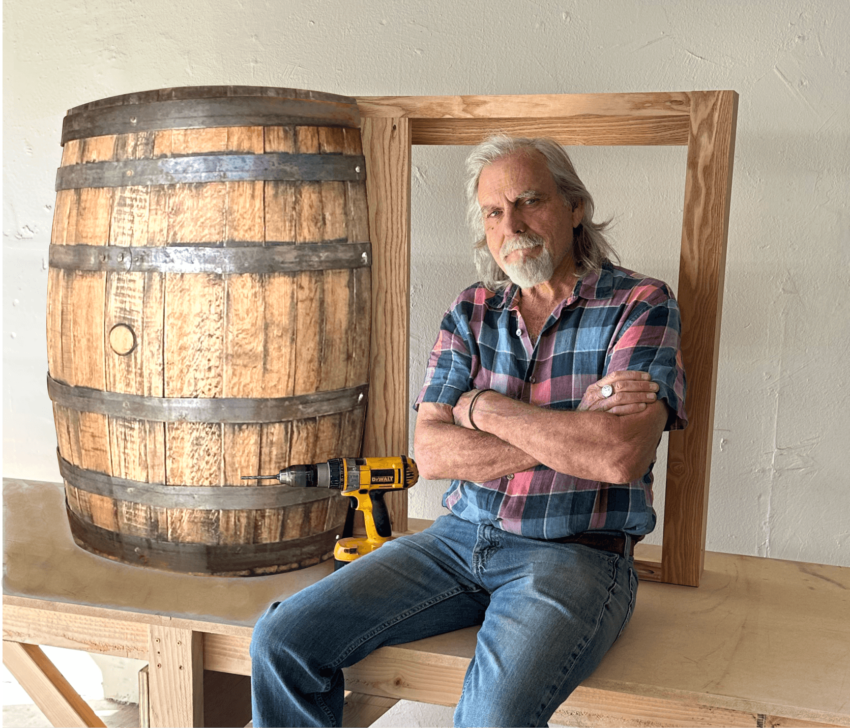 An older man with long gray hair and a beard, wearing a plaid shirt and jeans, sitting on a workbench. He has crossed arms and is seated next to a large wooden barrel with metal bands, in a room with white walls. There is a yellow power drill on the workbench in front of him.