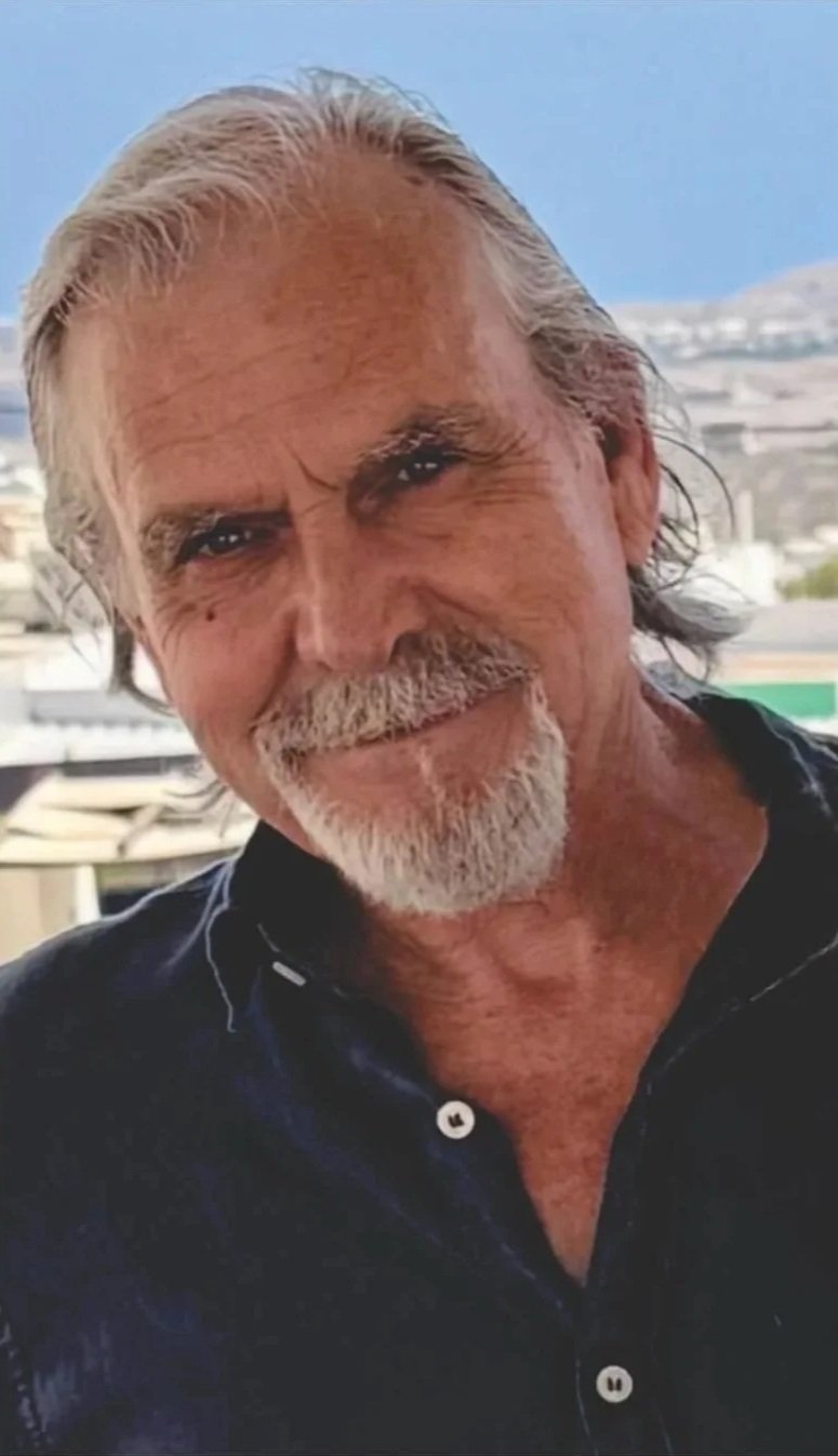 Close-up of an older man with gray hair, beard, and mustache, smiling outdoors with a blurred cityscape in the background.