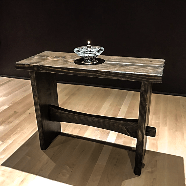 A rustic wooden table with a glass candle holder on top, placed on a wooden floor against a dark wall.