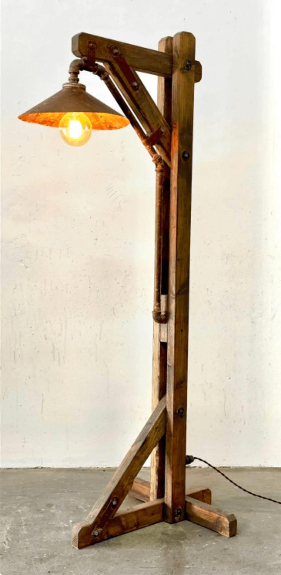 A rustic wooden floor lamp made of reclaimed wood with a single exposed light bulb and a metal shade, standing on a concrete floor against a plain wall.