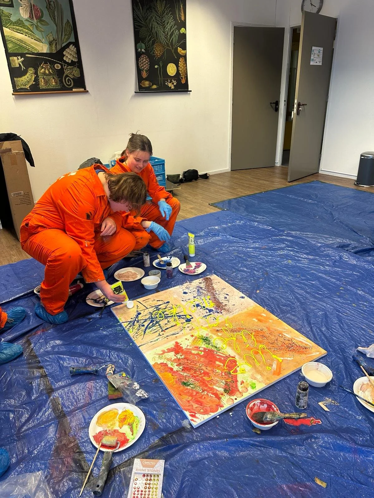 Twee mensen in oranje overalls maken samen een kleurrijk kunstwerk op een doek, dat ligt op een blauwe plastic tarp. Ze glimlachen en gebruiken verf en andere materialen in een interieurruimte.