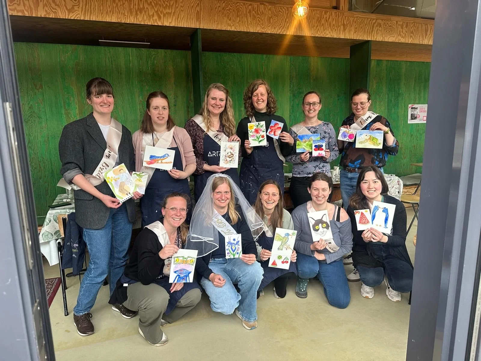 Een groep van twaalf vrouwen die in een kamer staan, sommigen zitten en anderen staan, allemaal houden ze kleurrijke kunstwerken in de hand, en één vrouw draagt een bruiloftsveil. Ze lijken blij en poseert voor de foto.