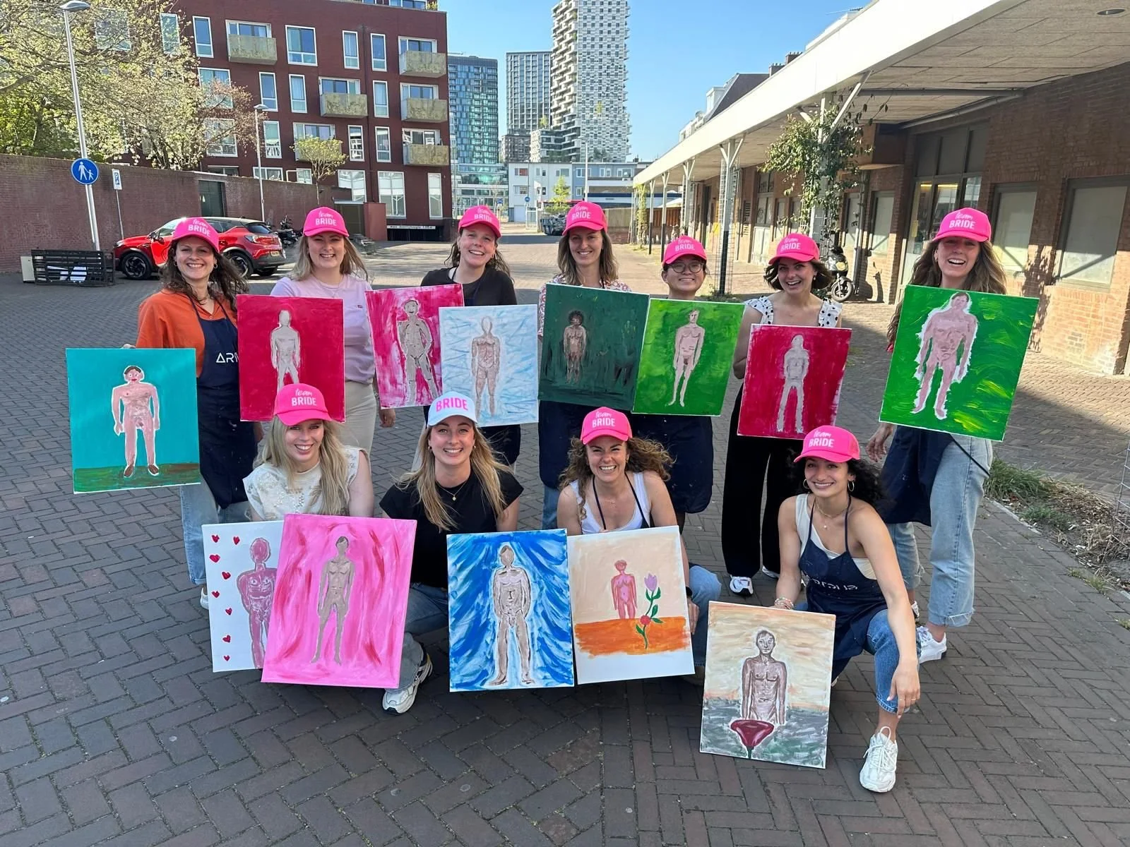 Groep vrouwen met roze petten met de tekst 'team BRIDE' die in een buitenruimte staan, terwijl ze schilderijen vasthouden van menselijke figuren in verschillende stijlen.