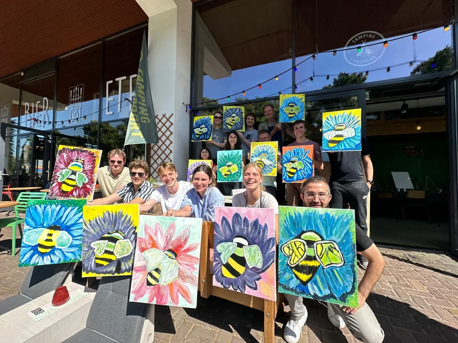 Groep mensen posed met kleurrijke schilderijen van bijen op een terras buiten.