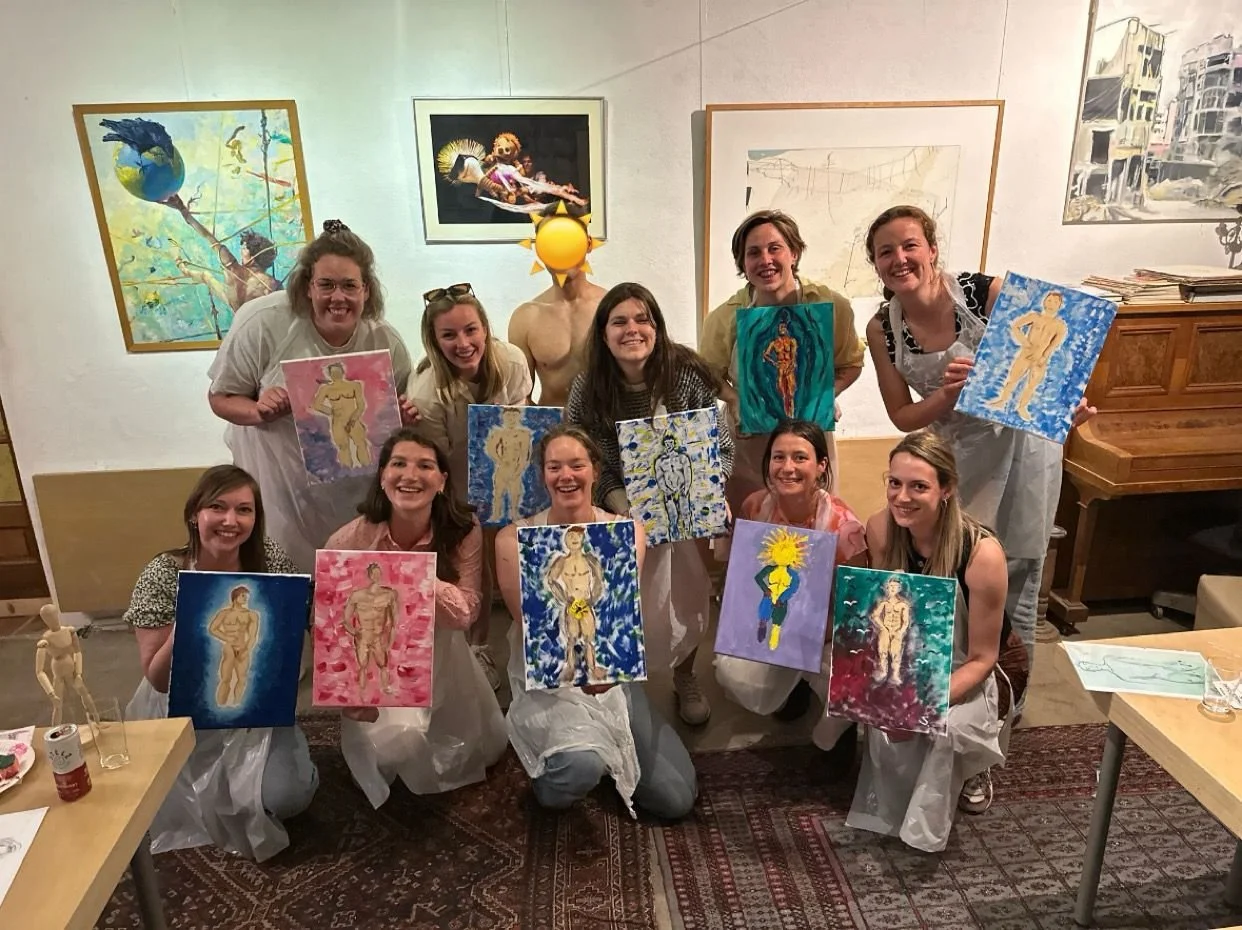 Vijftien mensen die deelnemen aan een schilderworkshop, sommige houdend hun kunstwerken, in een kunstgalerie. Ze lachen en poseren voor de foto, met een piano en kunstwerken op de muur op de achtergrond.