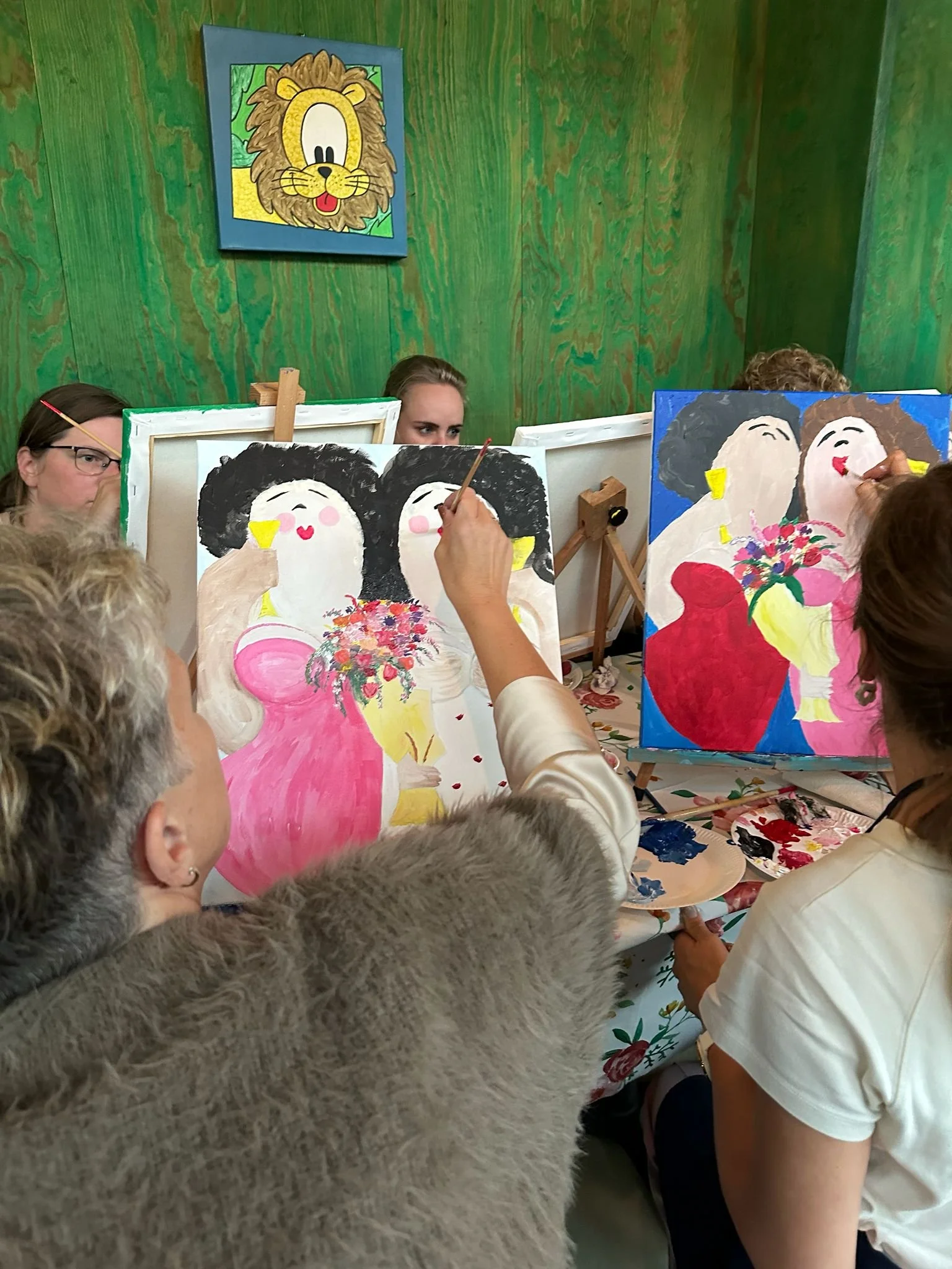 Mensen schilderen portretten van vrouwen met parrua, bloemen en kleurrijke kleding in een schilderworkshop, met groene houten muren en een cartoon van een leeuw op de muur.