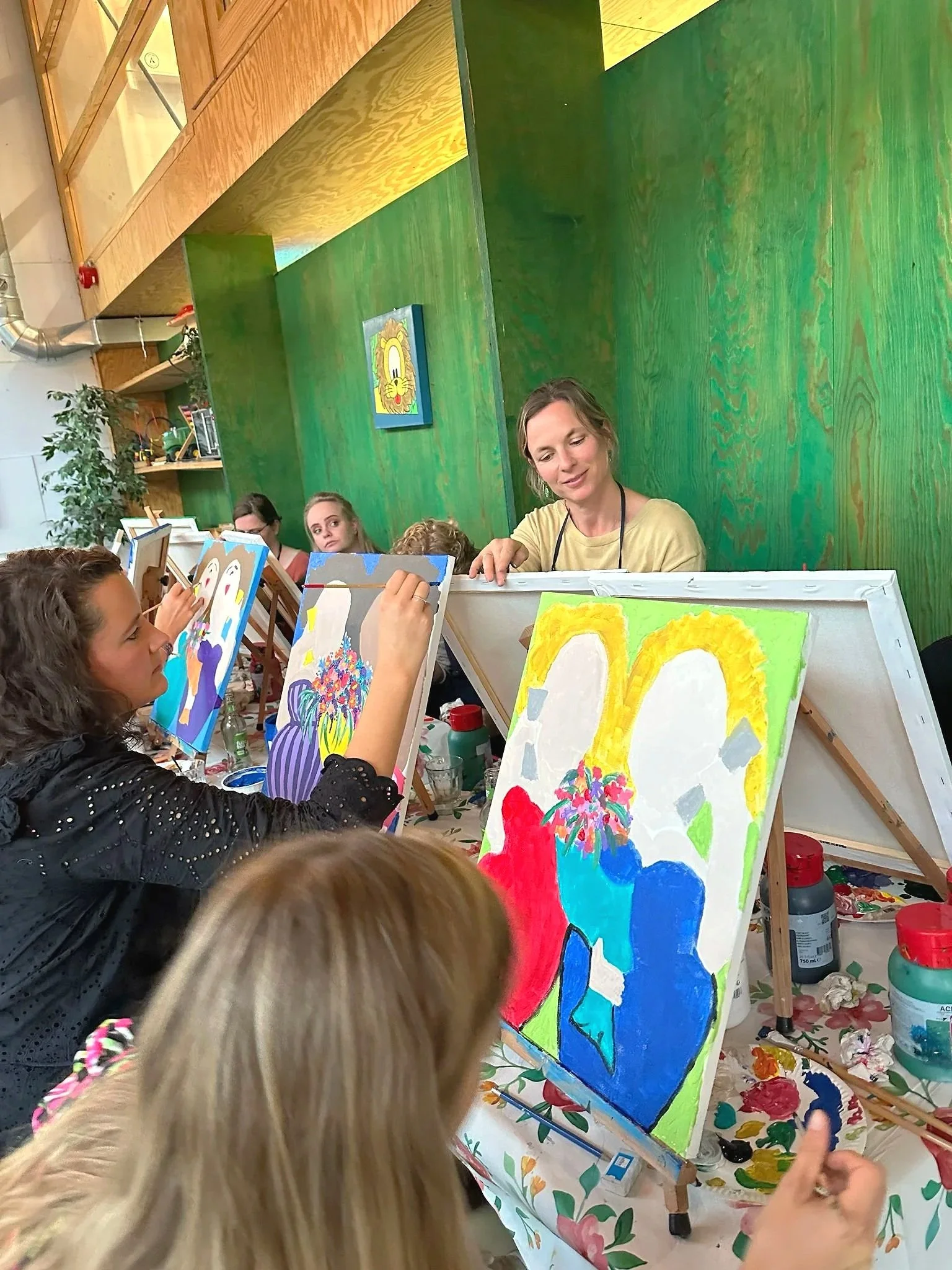 Een groep mensen die samen in een schilderworkshop aan het schilderen zijn, met kleurrijke schilderijen voor zich en een groene achtergrondmuur.