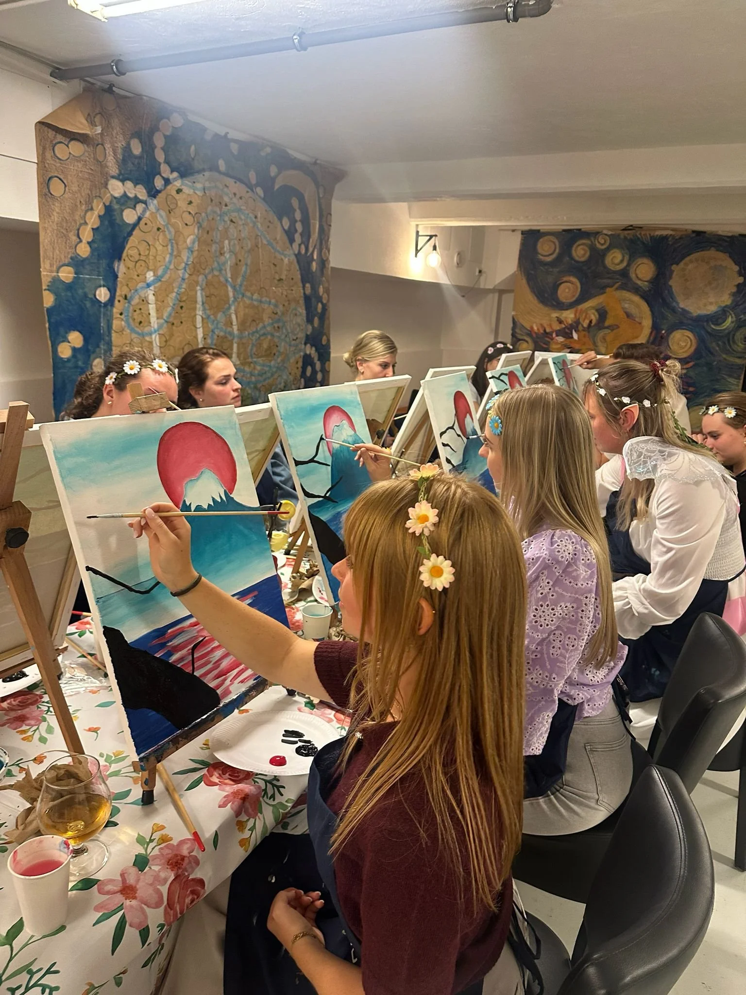 Kinderen en volwassenen die schilderen op canvas met een artistieke achtergrond in een kunststudio. Ze dragen bloemenkronen en richten zich op hun schilderijen.