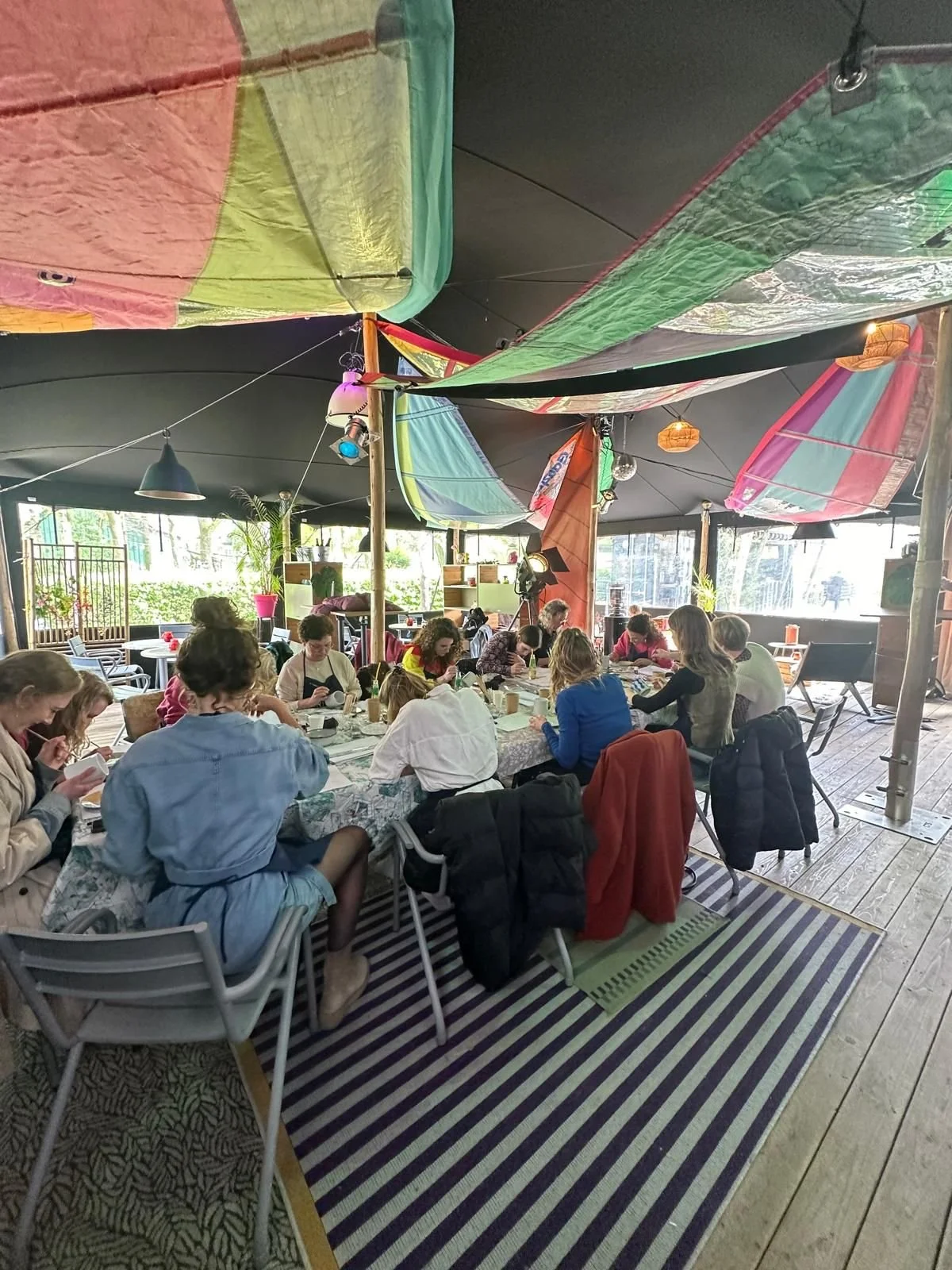 Een groep mensen zit gezamenlijk aan een tafel in een overdekt, kleurrijk interieur. Ze zijn bezig met een activiteit, mogelijk tekenen of schrijven, op een daglichtrijke locatie met decoraties en planten.