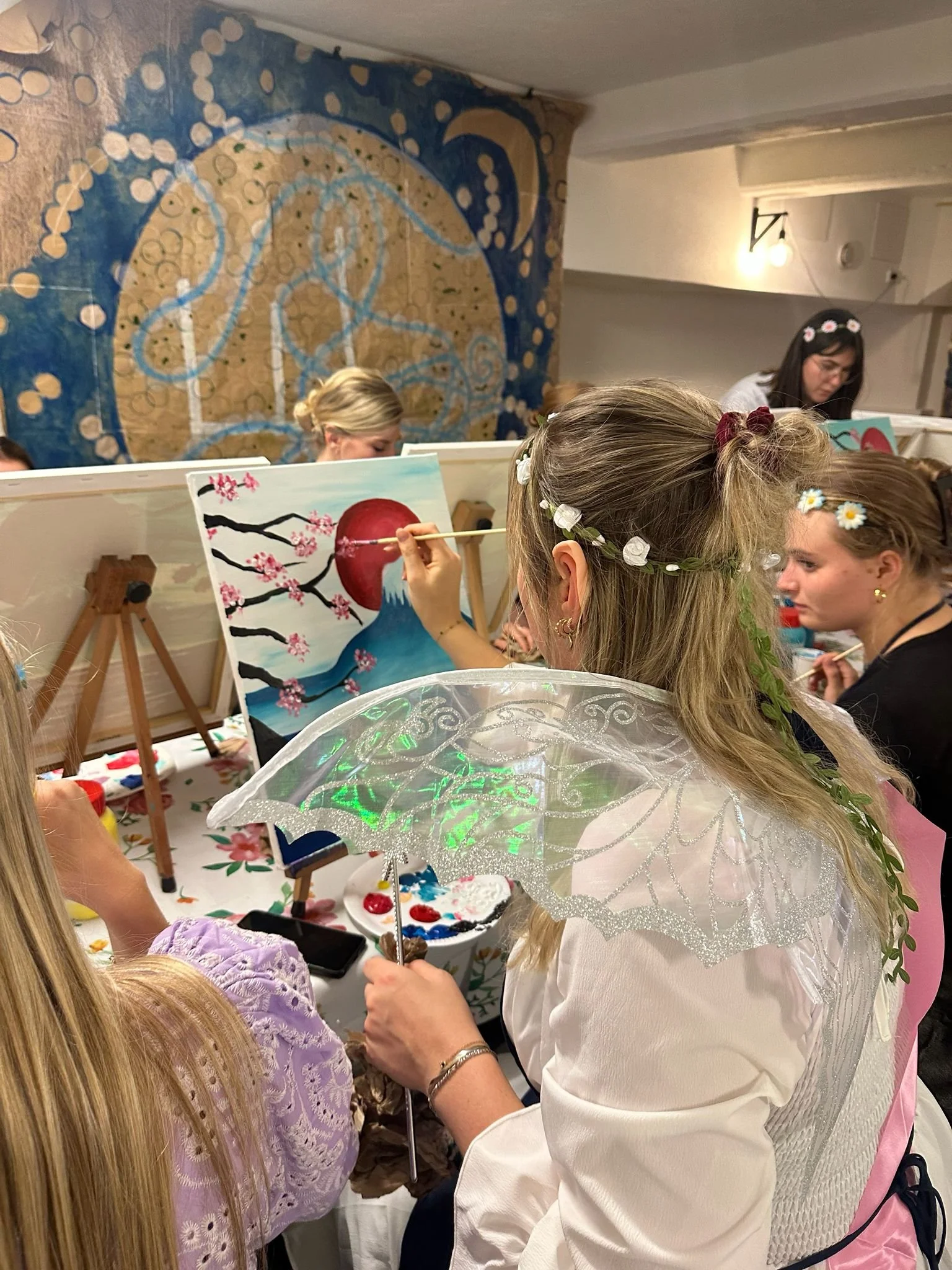 Een groep mensen, veel met bloemenkronen en lampions, die samen aan een schilderworkshop deelnemen in een gezellige ruimte. Ze schilderen kleurrijke kunstwerken, onder andere met thema's zoals een Japanse kersboom en een vulkaan met een rode zon.