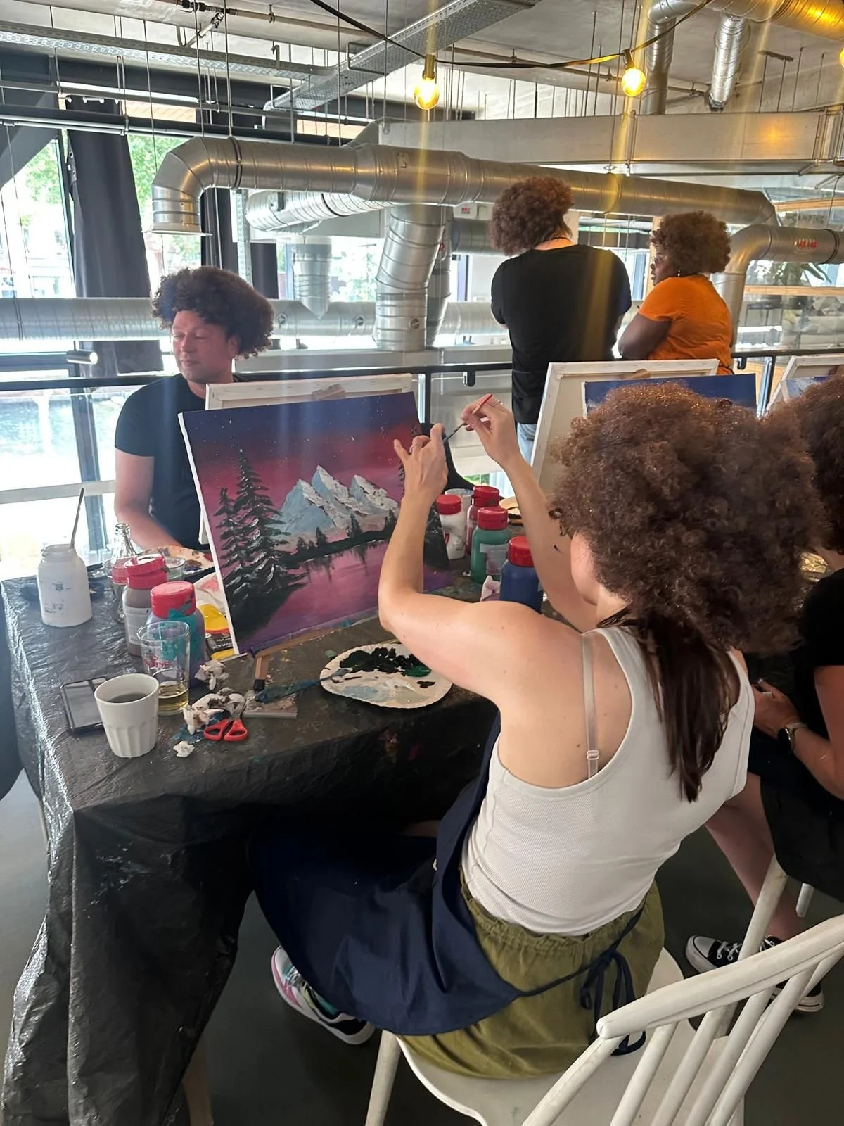 Mensen die samen een landschap schilderen, met bergen, bomen en een kleurrijke hemel op het canvas, in een moderne, industriale ruimte.