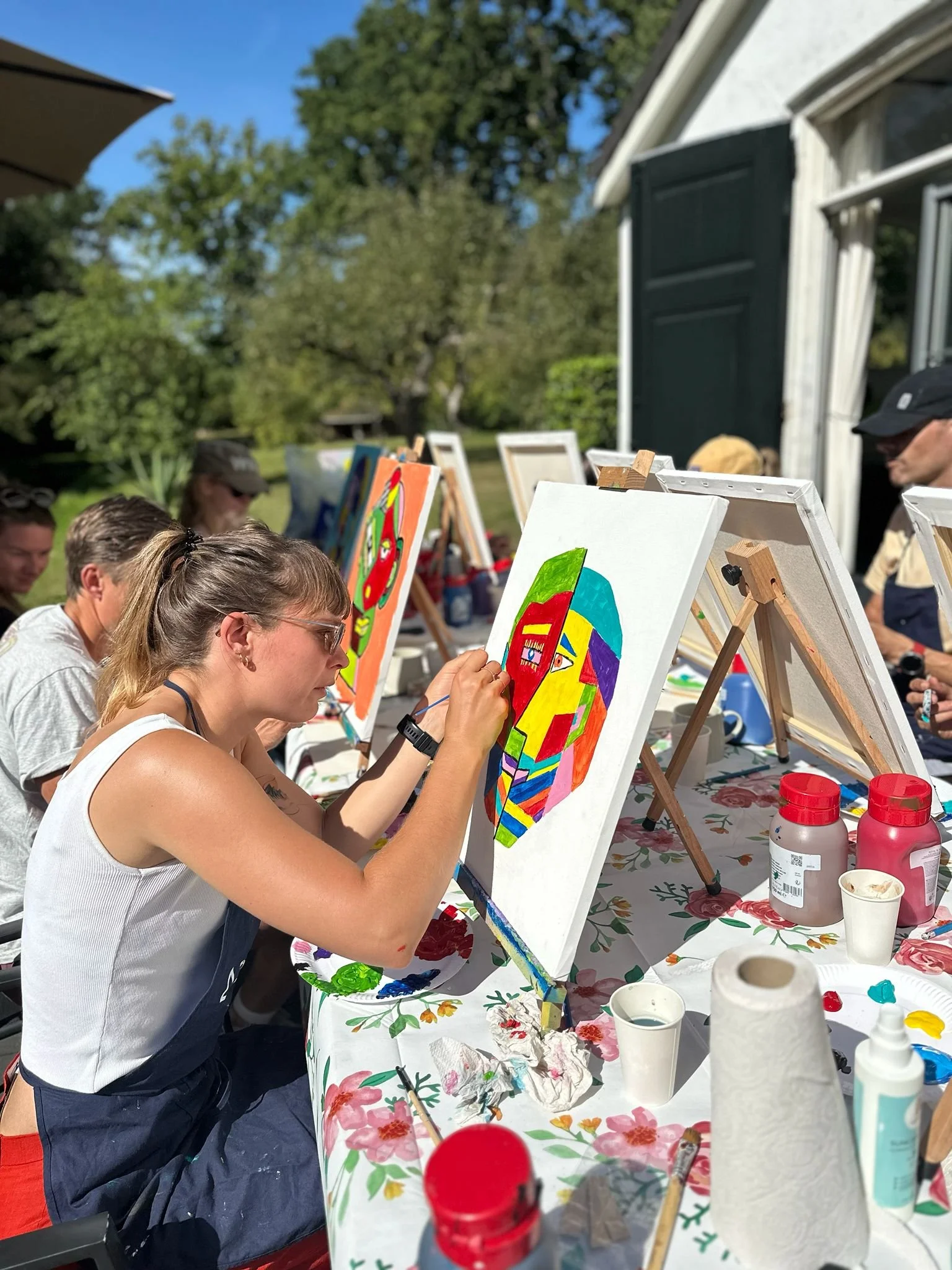 Mensen die buiten schilderen op een zonnige dag met kleurrijke kunstwerken op canvas, met groene bomen en een huis op de achtergrond.
