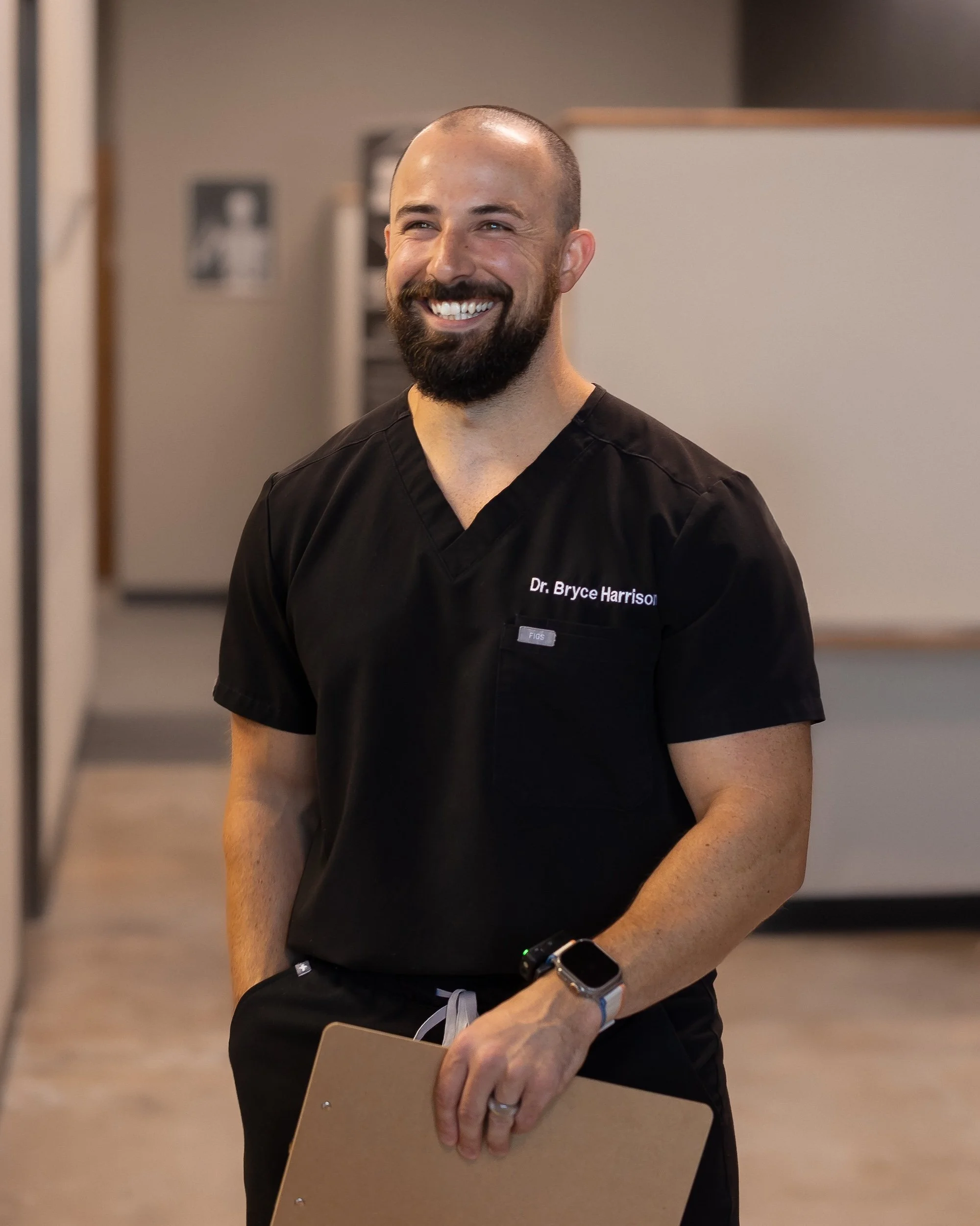 A man in medical scrubs holding a clipboard, smiling in a professional setting.