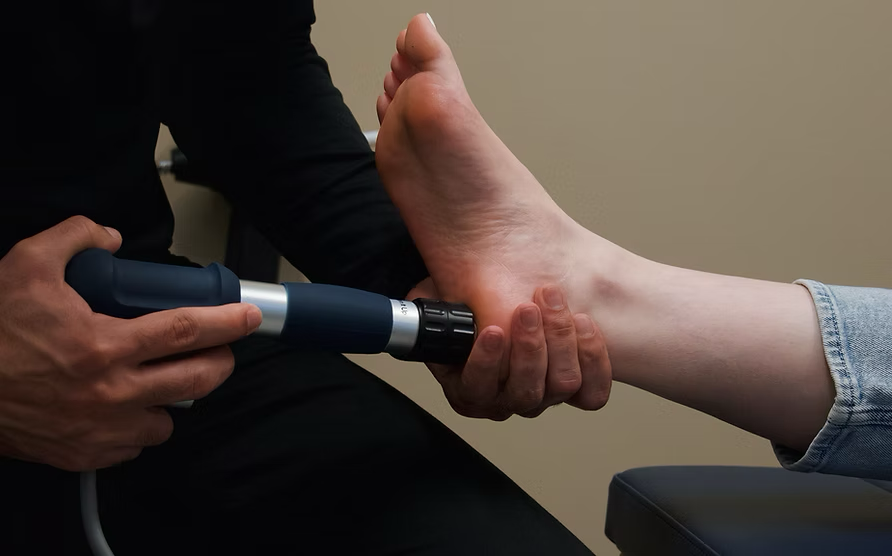 Shockwave Therapy. A person holding a foot with one hand and using a medical device on the sole of the foot with the other hand.