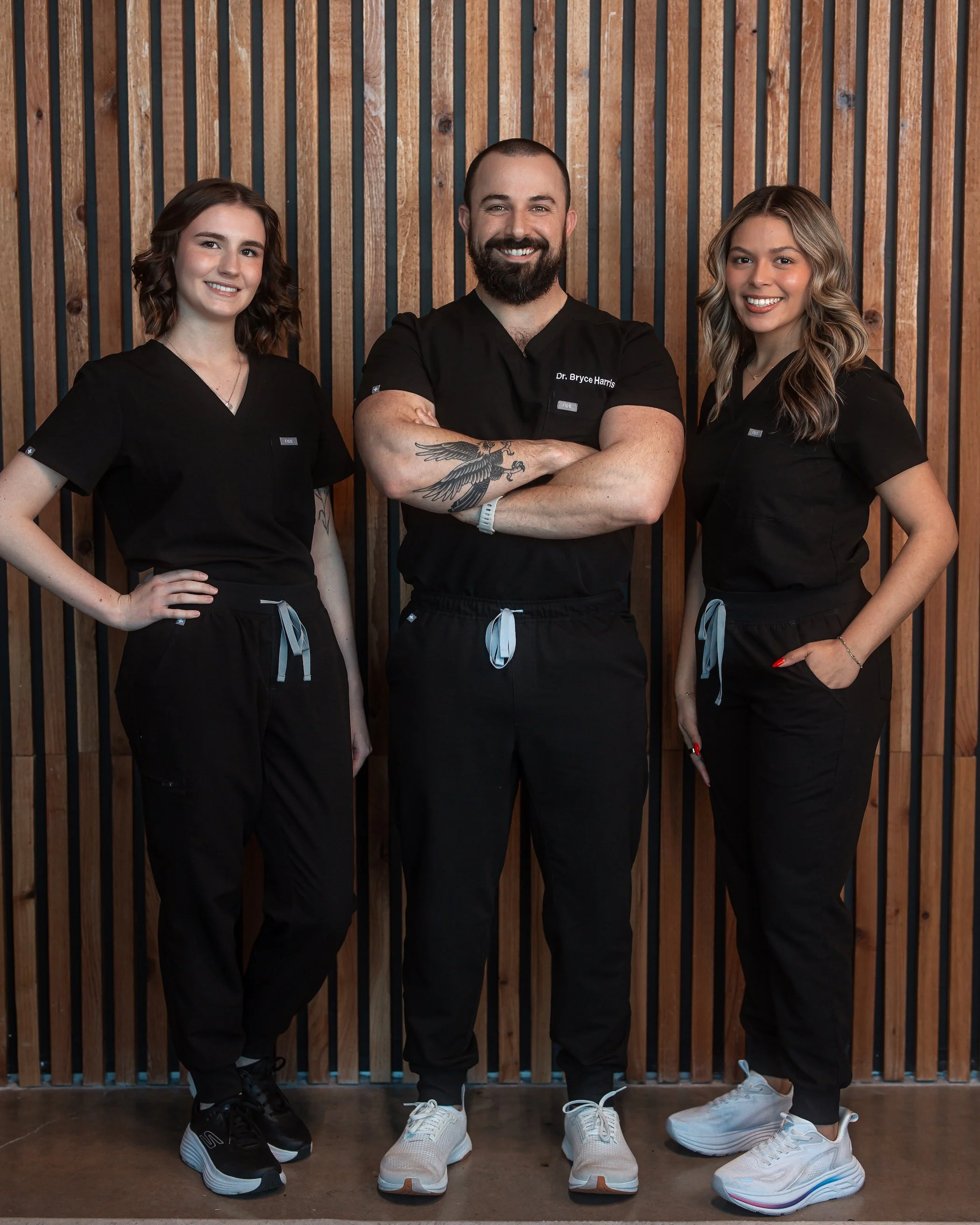 Three healthcare professionals standing in front of a wooden slat wall, smiling at the camera. The man in the middle has a beard, tattoos, and is wearing a black scrubs with his arms crossed. The two women on either side are also in black scrubs, with one on the left having short brown hair and the other on the right having wavy blonde hair.