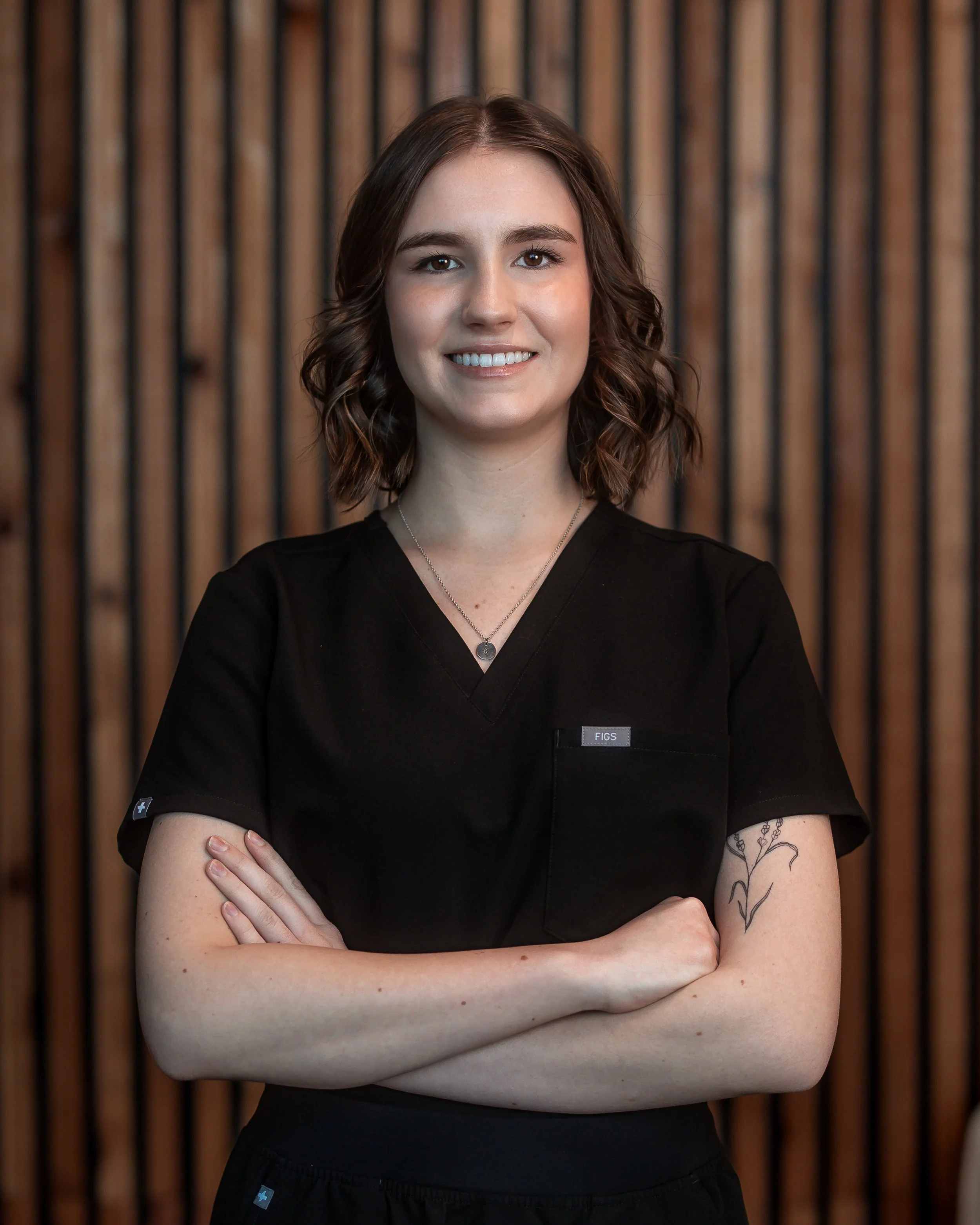 A young woman with shoulder-length brown hair, wearing a black medical uniform, standing with arms crossed in front of a wooden wall background.