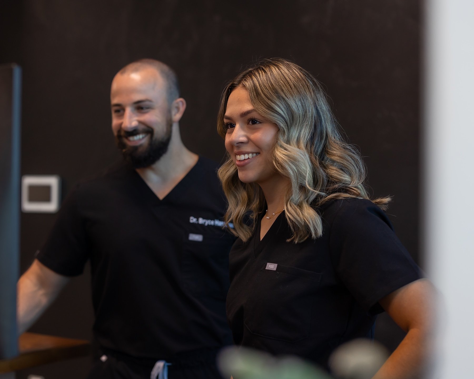Two healthcare professionals, a man with a beard and a woman with wavy blond hair, smiling and wearing black scrubs, standing indoors against a dark background.