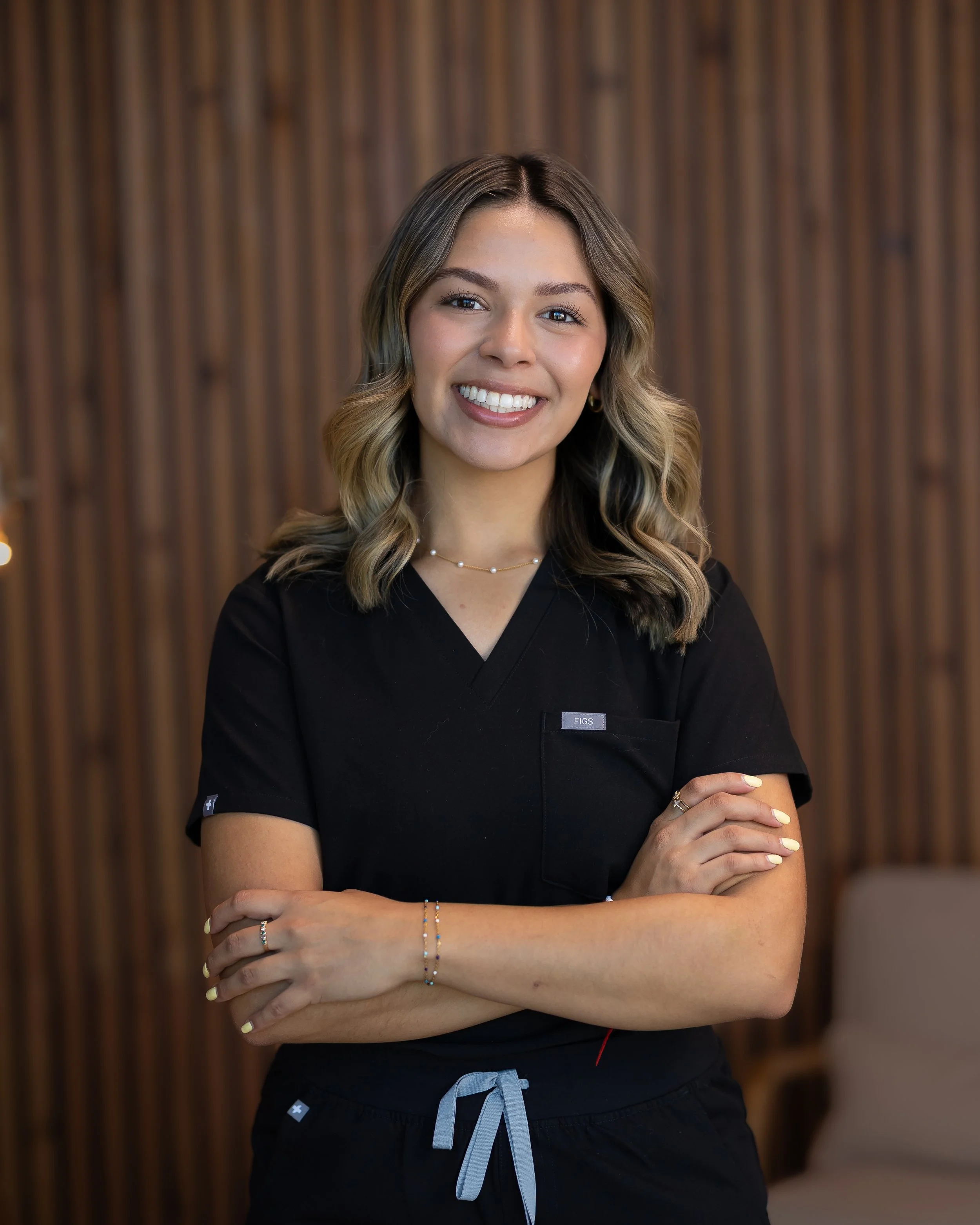 A smiling woman with wavy hair wearing black scrubs, standing with arms crossed indoors, with a wooden wall background.