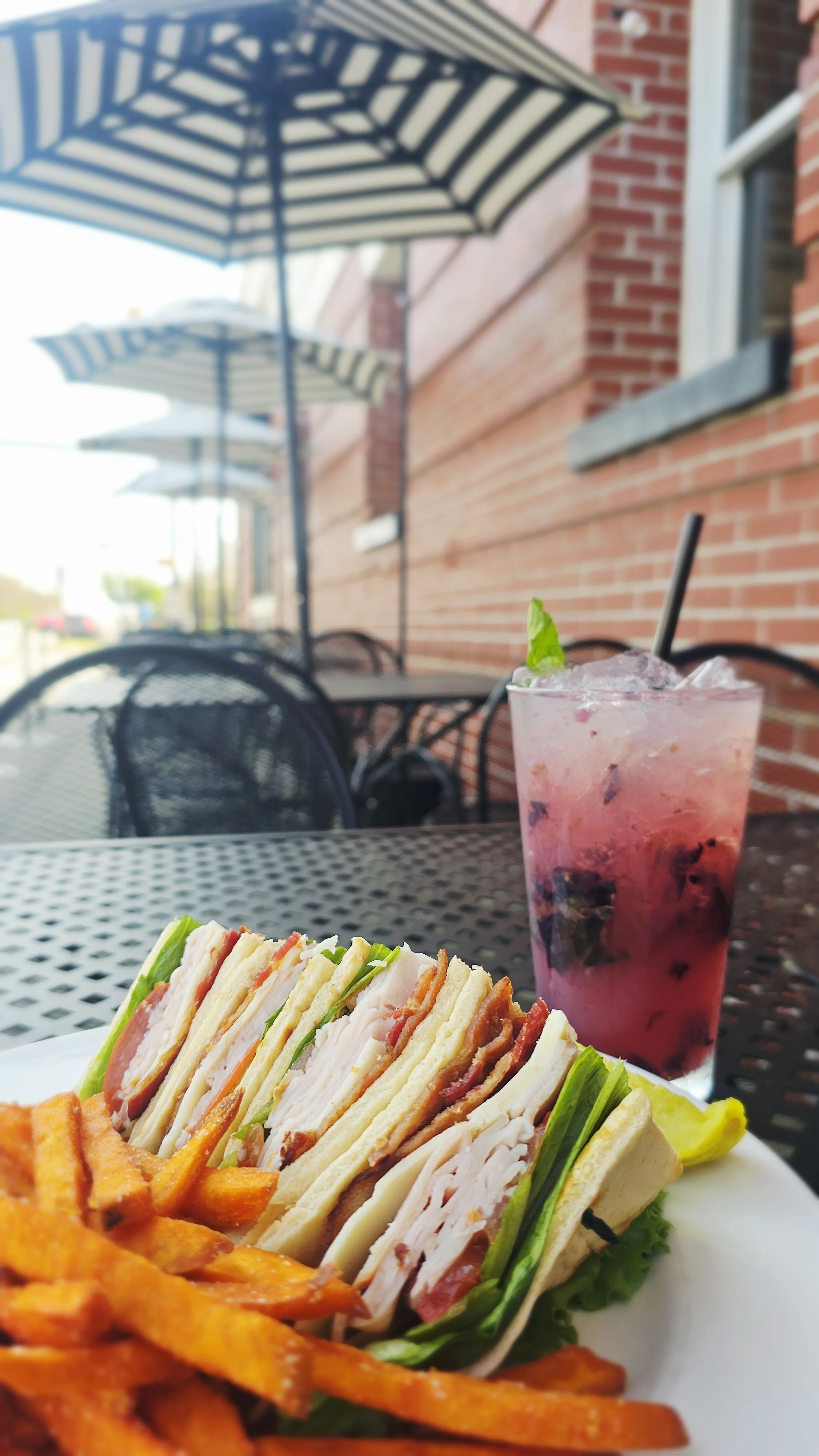 A plate with stacked sandwiches, a side of sweet potato fries, a pink berry drink with mint garnish, and french fries on a black metal outdoor table, with striped umbrellas and a red brick building in the background.