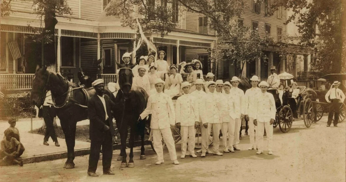 Historical black and white photo of a large group of people dressed in early 20th-century clothing, some on horse-drawn carriages, gathered in front of a large house with trees around.