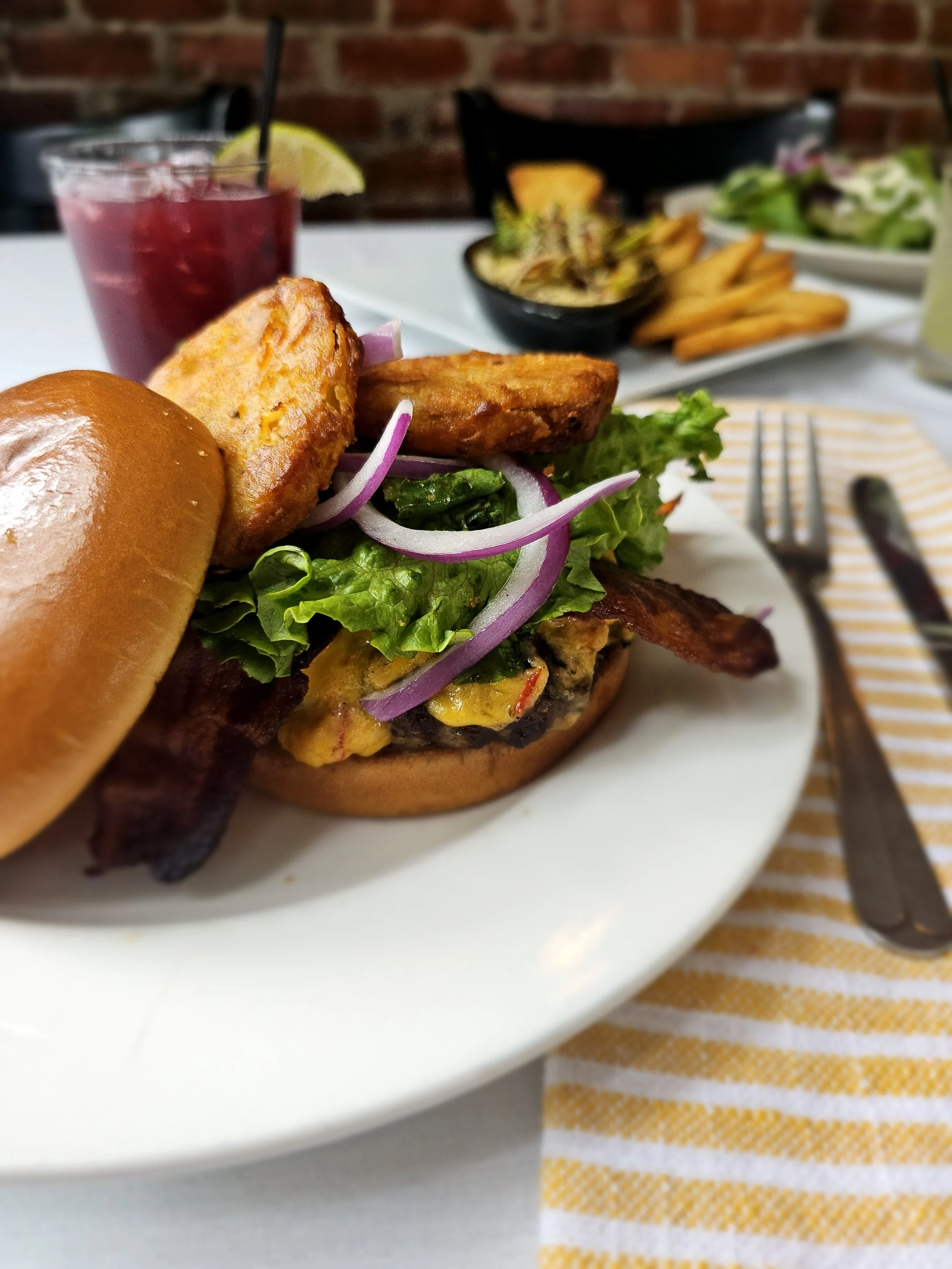 Close-up of a cheeseburger with leaf lettuce, tomato, onion slices, and two fried items, served on a bun. In the background, a glass of red berry drink with a lemon wedge and black straw, along with side dishes of salad, fries, and roasted vegetables on a white plate.