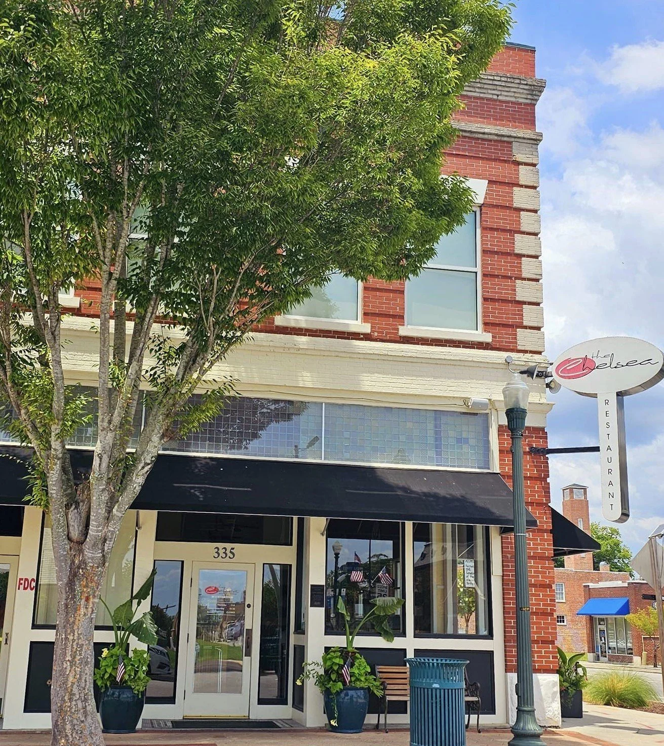 Exterior of a brick building with a restaurant entrance, black awning, and large windows, with a tree in front and a sidewalk in an urban area.