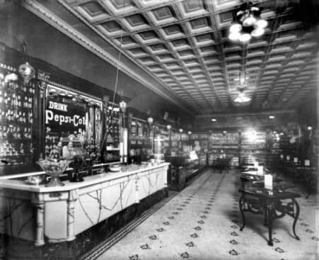 Historic black and white image of an old-fashioned bar with a long marble counter, vintage furniture, and ornate ceiling details.