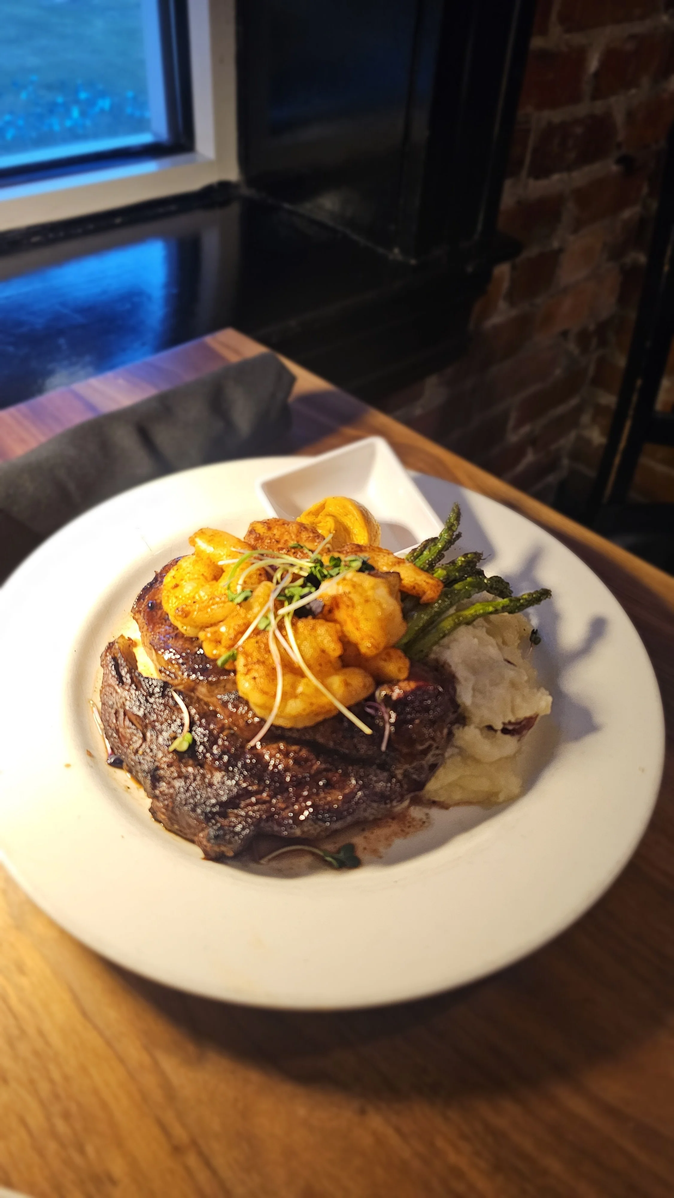 A plate with a cooked steak topped with grilled vegetables, mashed potatoes, and a small bowl of sauce, on a wooden table in a restaurant setting.