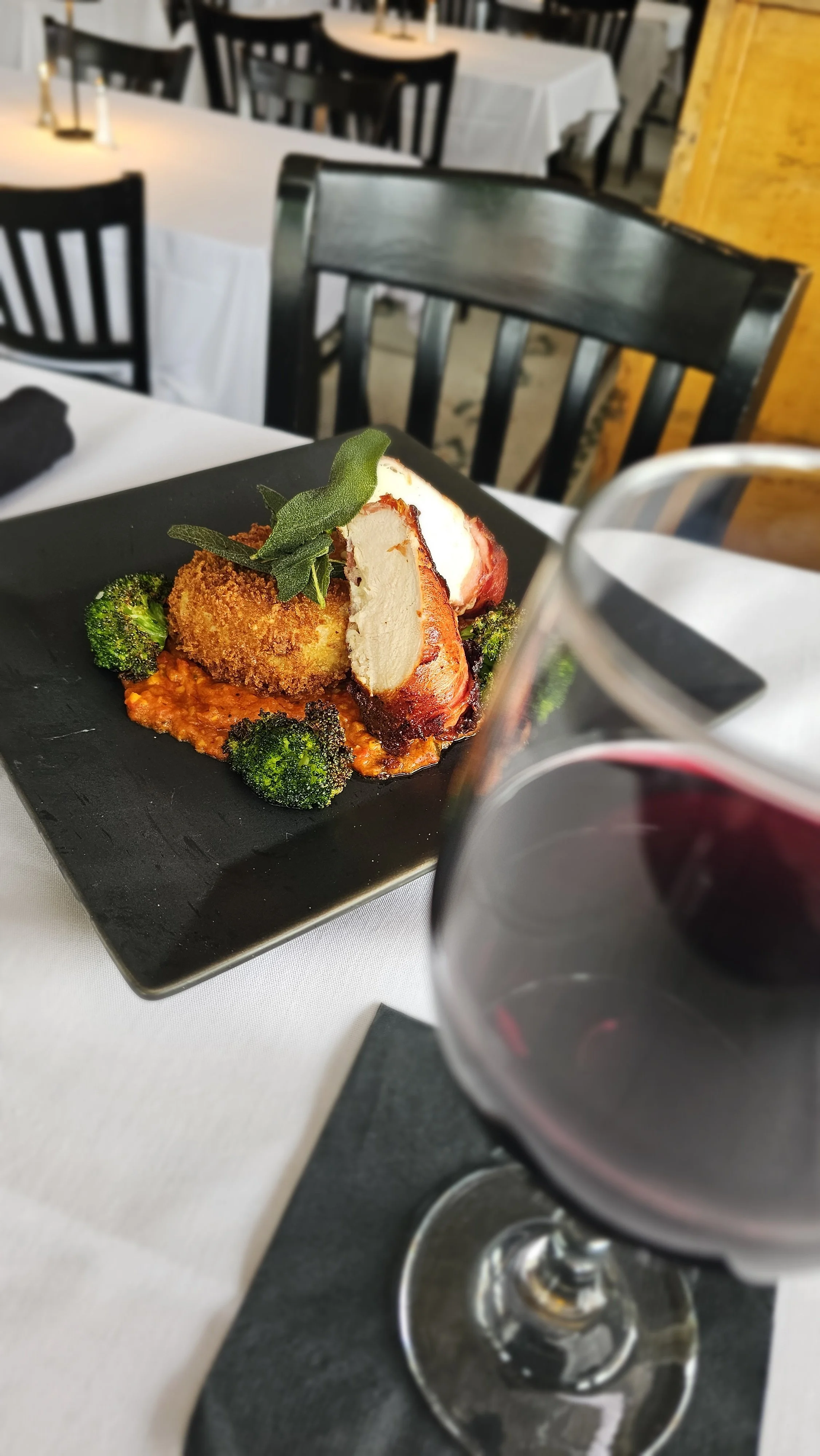 A plate with breaded fried chicken, roasted potato, broccoli, and garnished with sage leaves on a black square plate, with a glass of red wine in the foreground, set on a white tablecloth in a restaurant with black chairs and tables in the background.