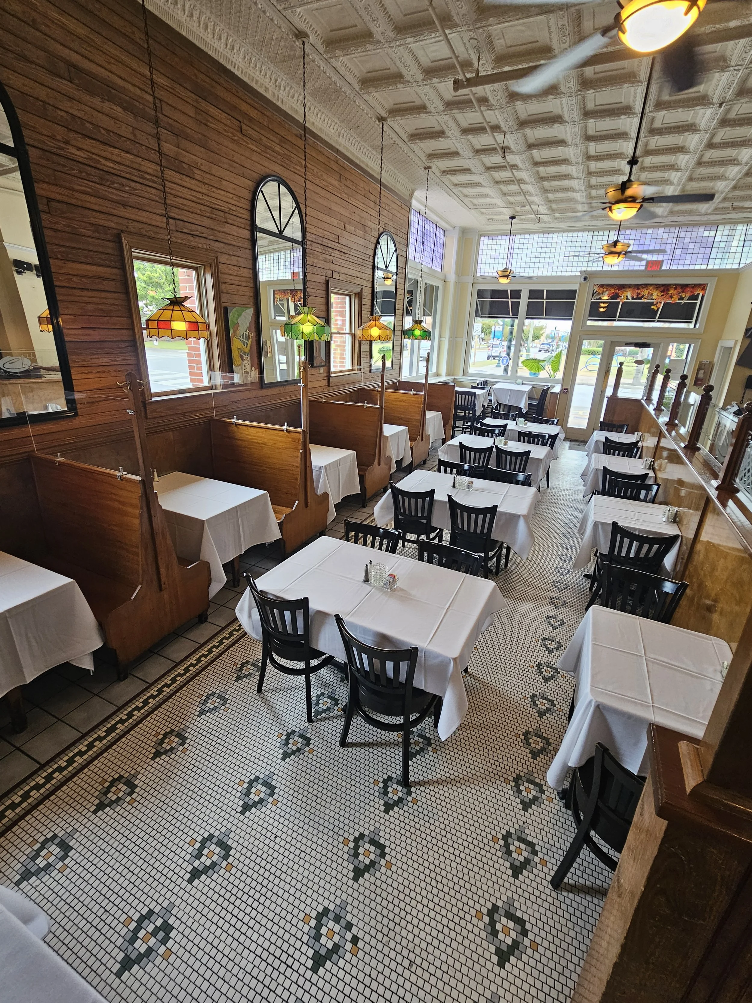 Empty restaurant with white tablecloth-covered tables, black chairs, decorative tile flooring, wooden booth seating, stained glass hanging lamps, and large windows allowing natural light.