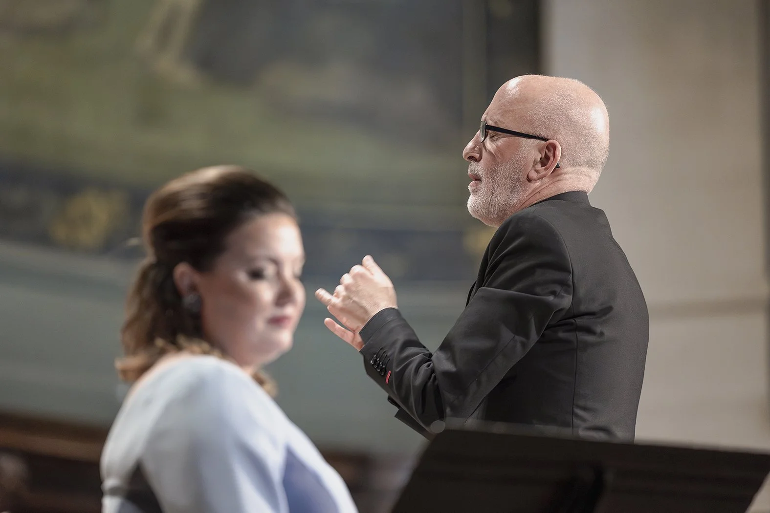 Hombre calvo, de gafas, vestido con saco negro, hablando con mujer con cabello rizado y vestido blanco en interior.