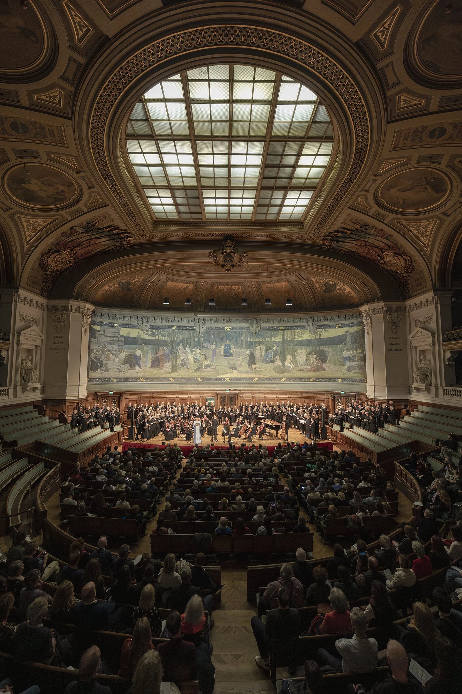 Concierto de orquesta en un auditorio elegante con techos decorados y una gran pintura en la pared del escenario