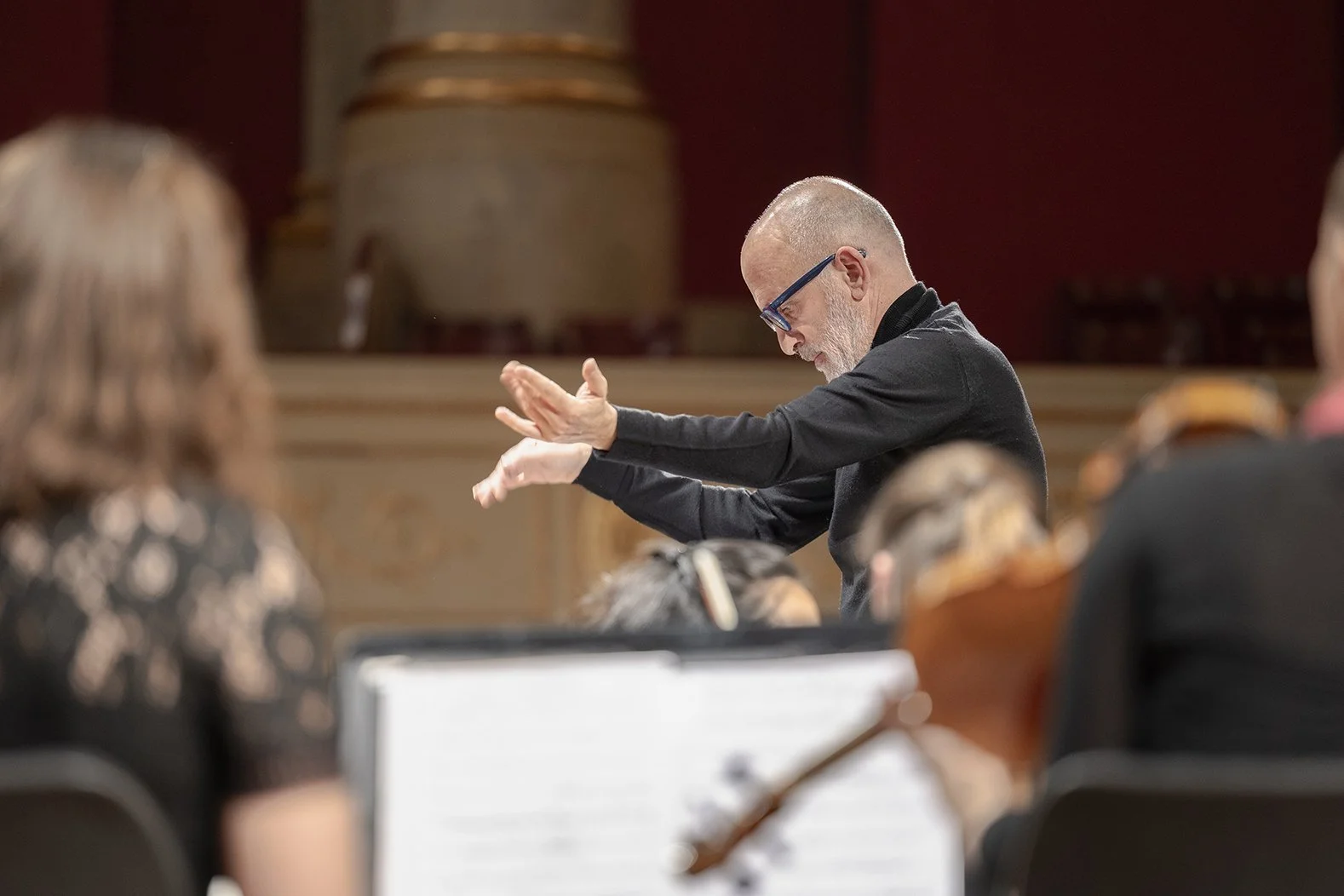 Un director de orquesta dirigiendo una orquesta en un auditorio. La escena muestra a varias personas tocando instrumentos y el director con gestos de conducción.
