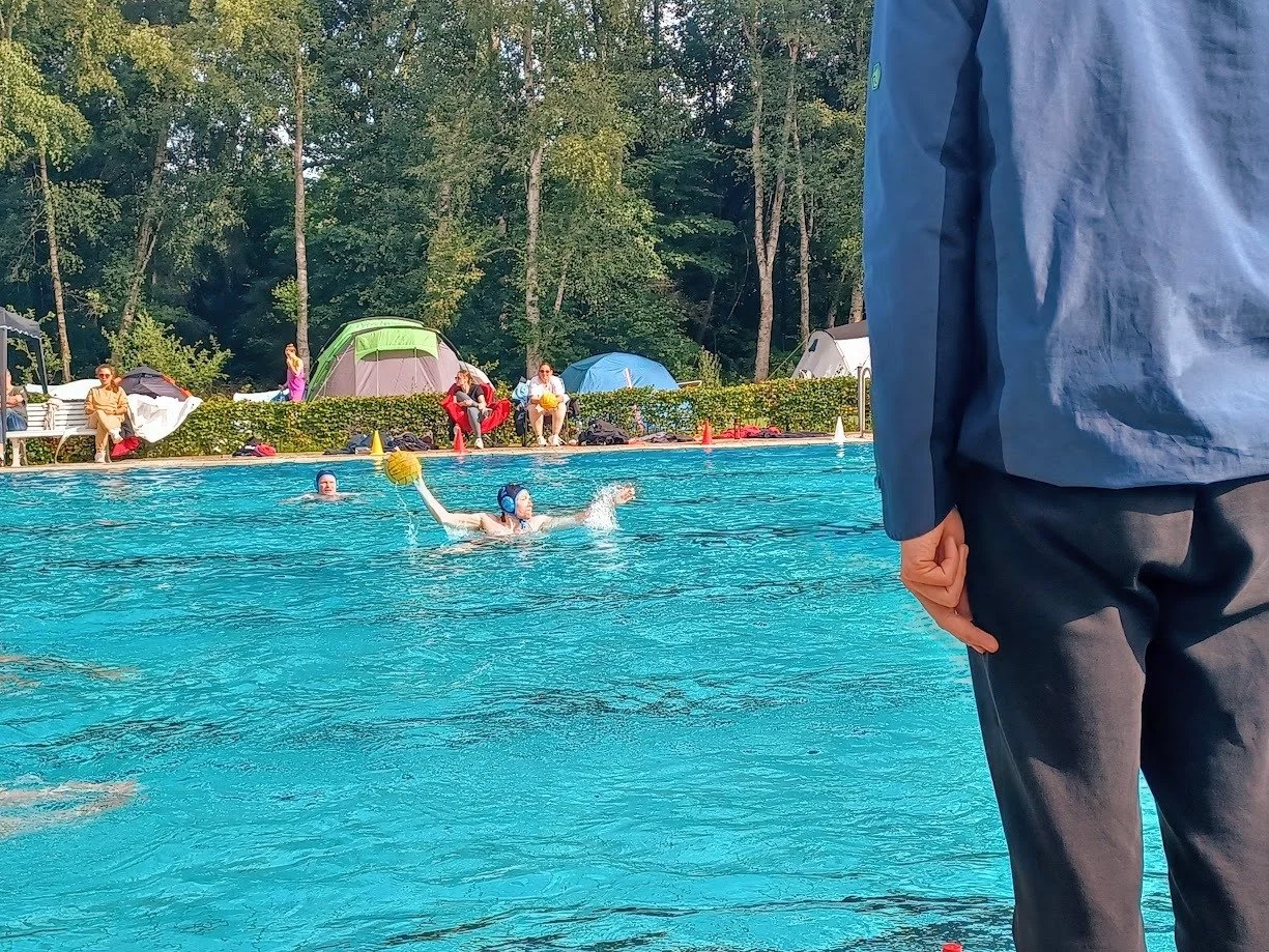 A person in a blue shirt observes a water polo game at an outdoor pool, with players swimming and reaching for a yellow ball in the water, surrounded by tents and people sitting on the poolside.