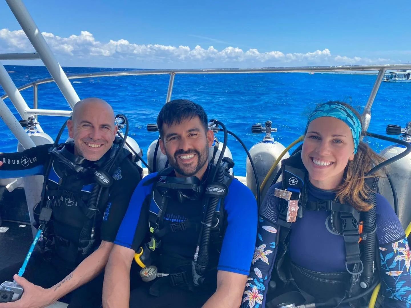 Three friends in scuba gear smiling on a boat with clear blue ocean and sky in the background.  Cozumel Scuba Diving Dive with k8