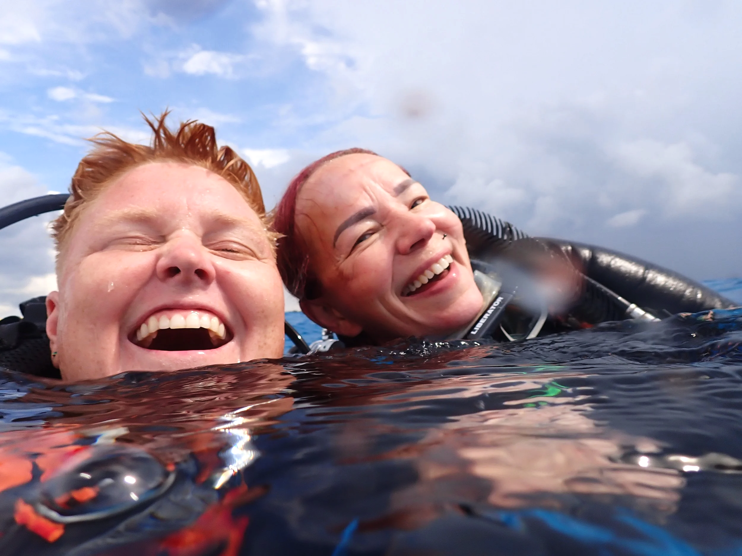 Two women smiling while swimming in the ocean, with water droplets on their faces and a partly cloudy sky in the background.  Cozumel Scuba Diving Dive with k8