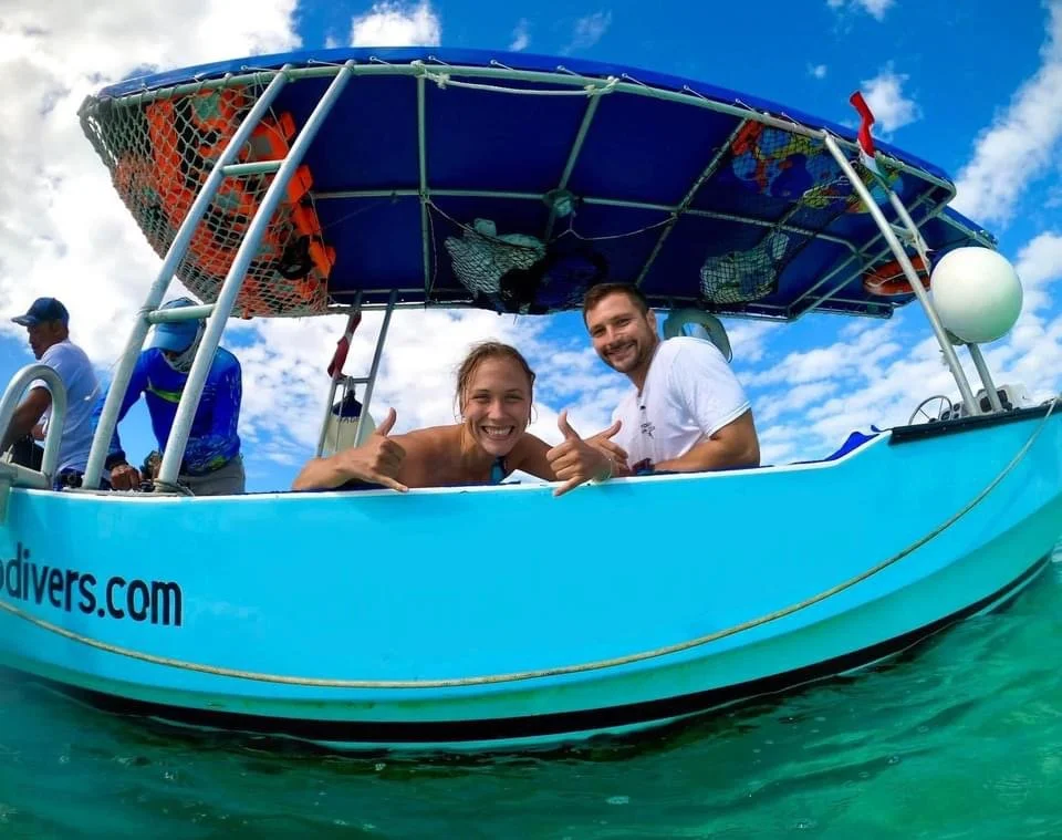 Two smiling people on a boat giving thumbs up, with a man at the helm and a woman waving. The boat is on clear water with a partly cloudy sky.  Cozumel Scuba Diving Dive with k8