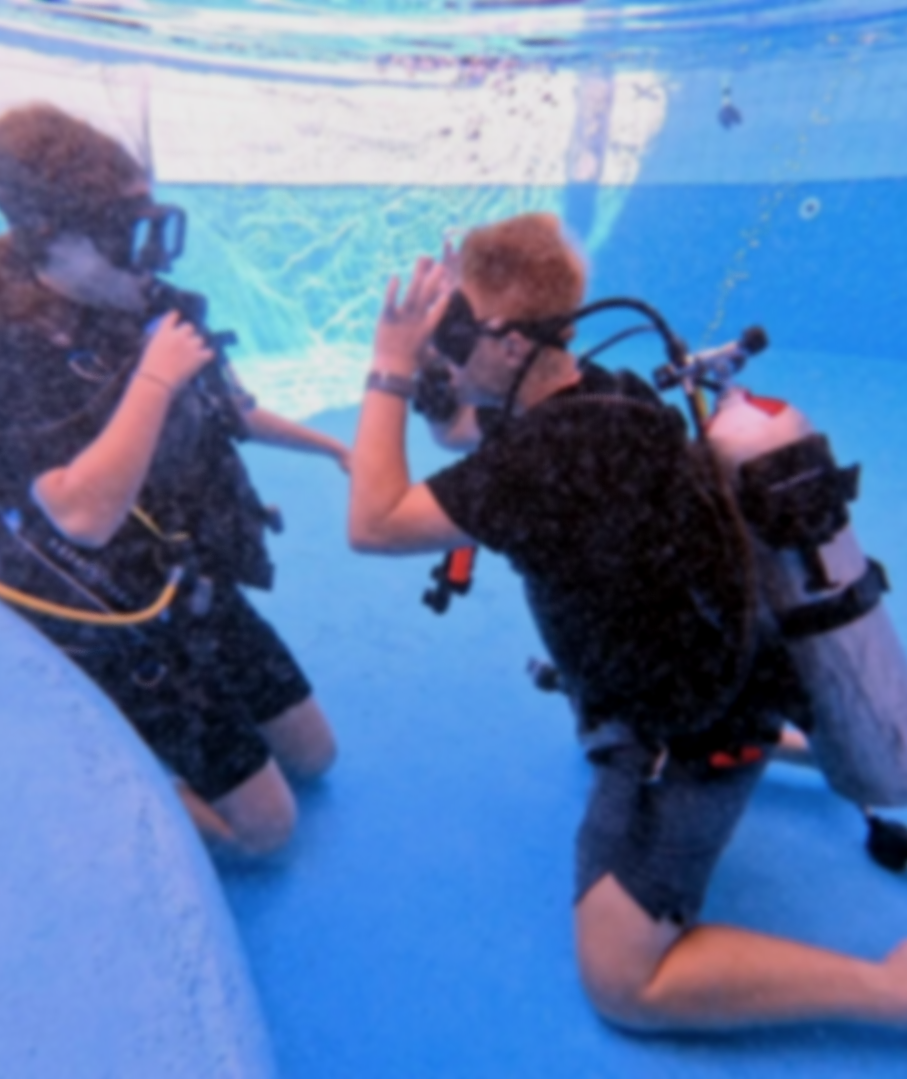 Two young boys are in a swimming pool, wearing scuba gear and engaging with a woman who is also underwater, all participating in a scuba diving activity.  Cozumel Scuba Diving Dive with k8