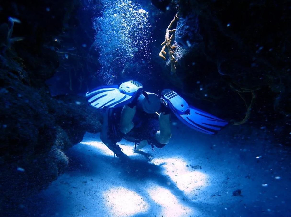 A person scuba diving through a narrow underwater rock passage with rocky walls, wearing a wetsuit and flippers, illuminated by blue light.  Cozumel Scuba Diving Dive with k8