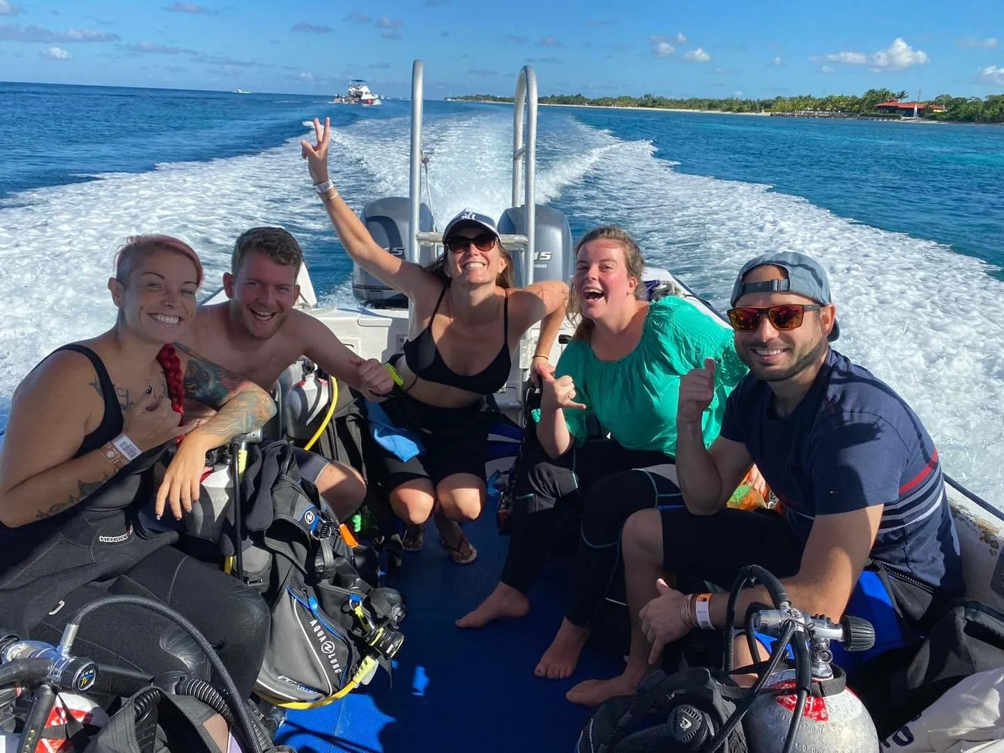Group of five smiling people on a boat, some with scuba gear, enjoying sunny weather on the water with distant shoreline and blue sky.  Cozumel Scuba Diving Dive with k8