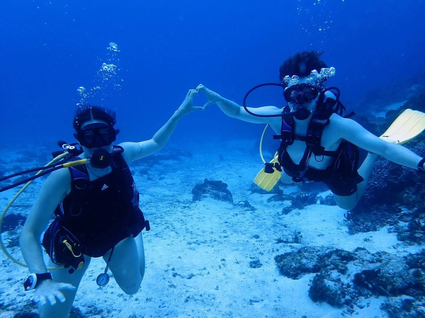 Two scuba divers underwater forming a heart shape with their hands, surrounded by blue water and coral.  Cozumel Scuba Diving Dive with k8