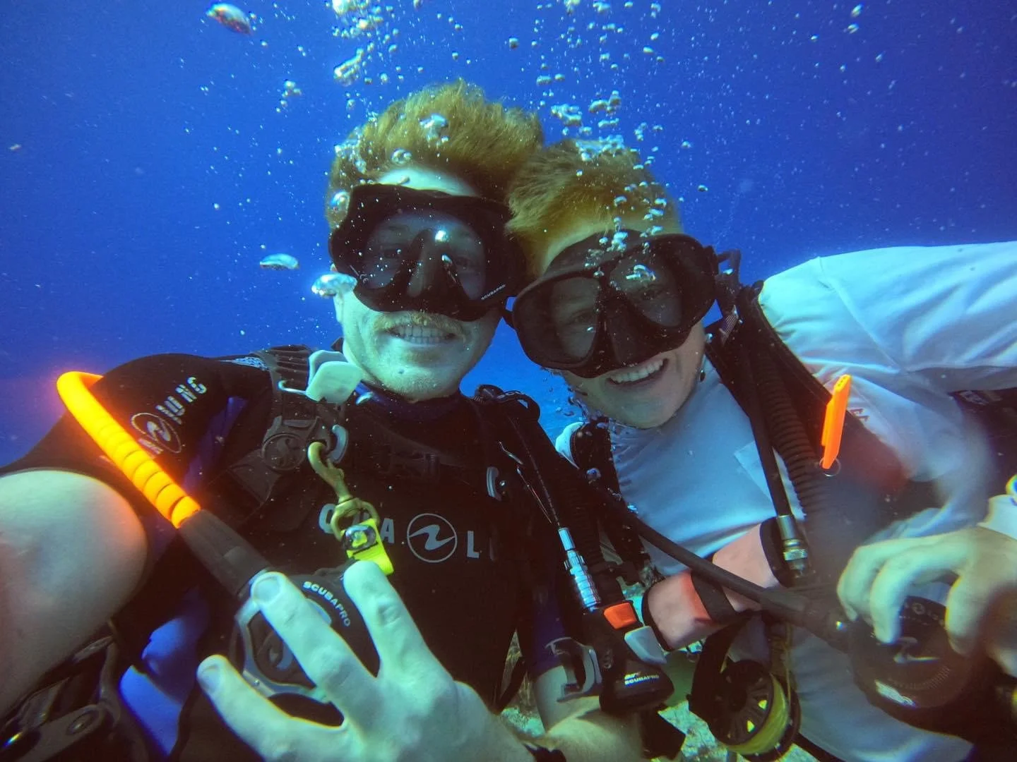 Two scuba divers underwater taking a selfie, smiling, with bubbles around them, wearing diving masks and wetsuits.  Cozumel Scuba Diving Dive with k8